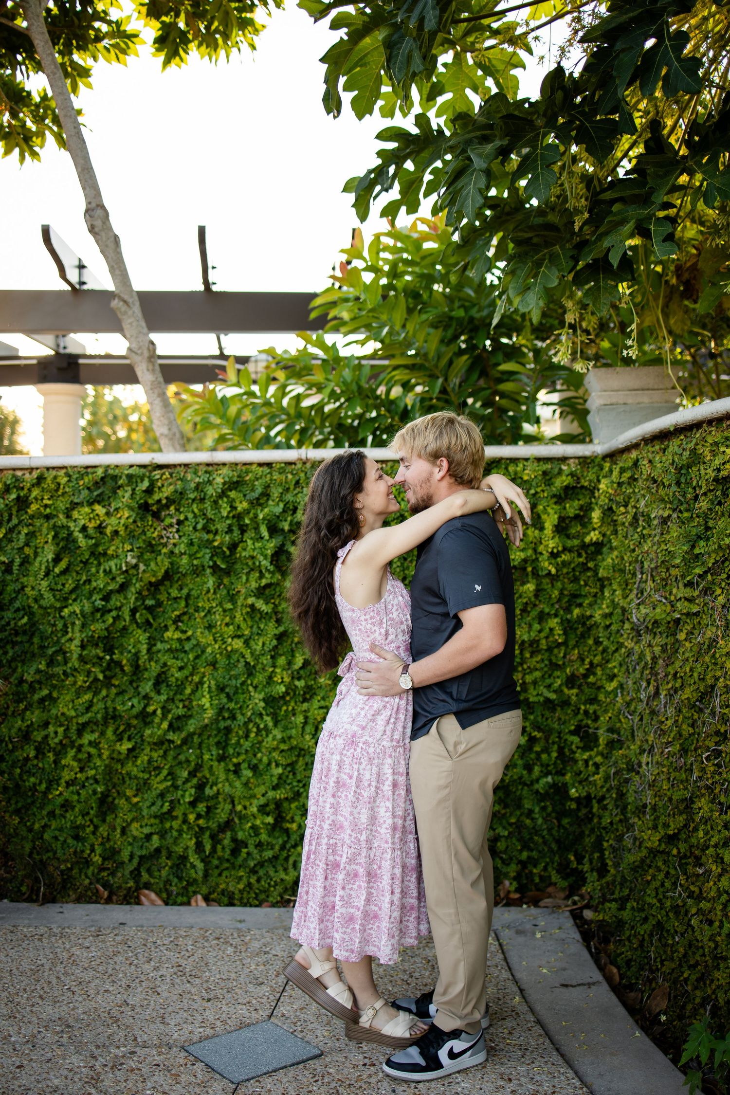 A couple shares a romantic kiss in front of a lush green hedge wall during a sunny day.