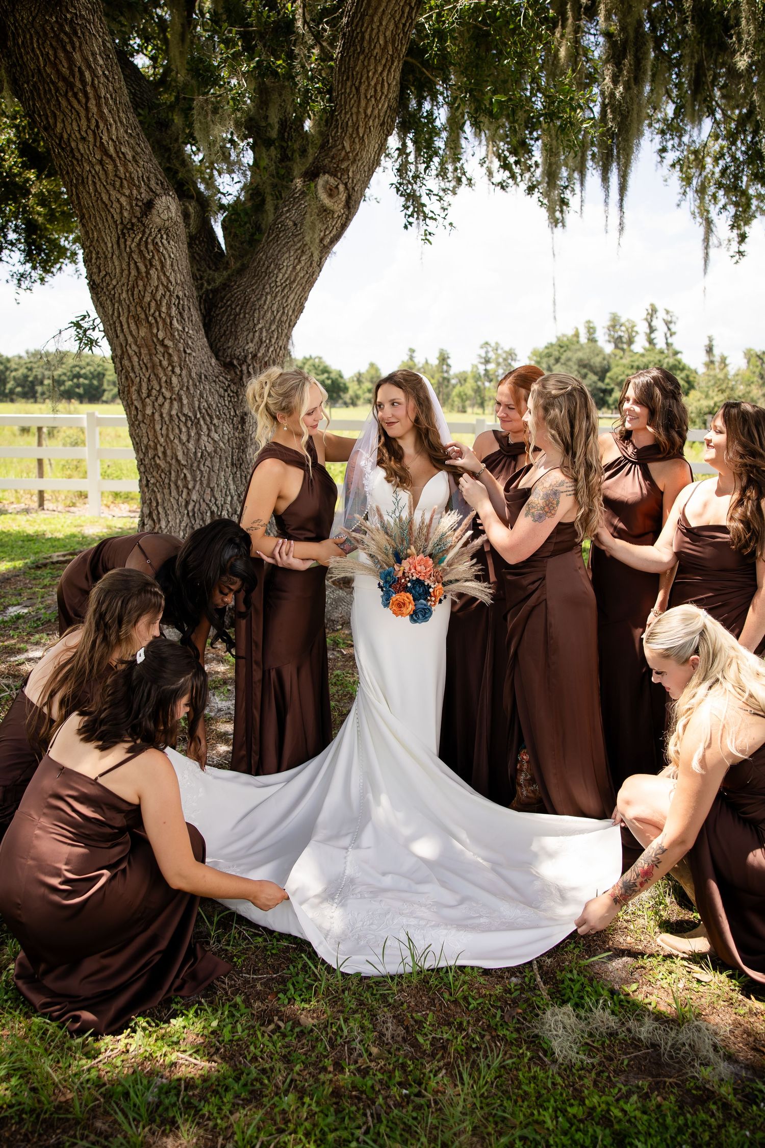 Bride in white gown holds colorful bouquet surrounded by bridesmaids in brown dresses under a large oak tree.