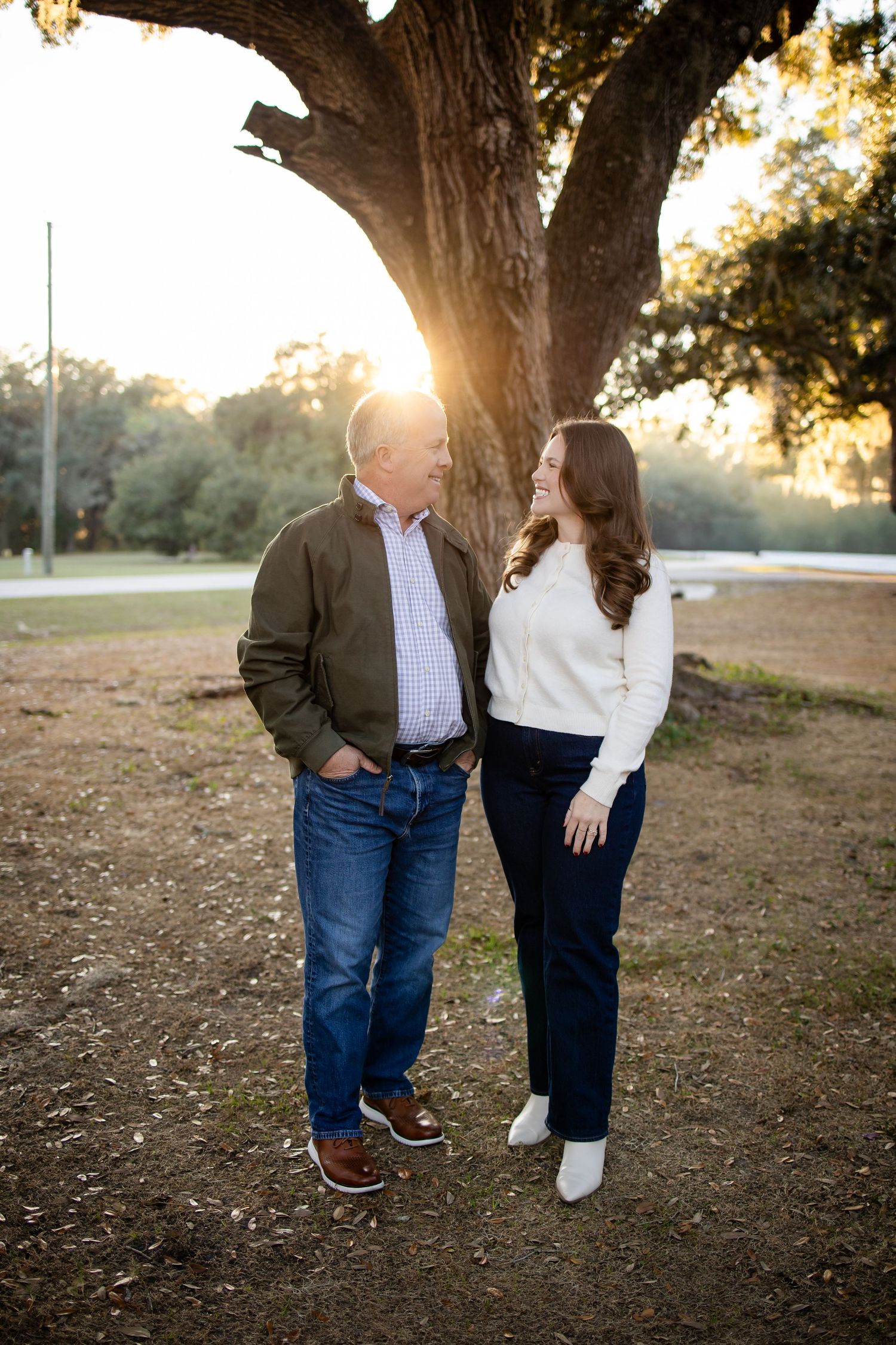Two people in casual attire stand together under a large tree at sunset.