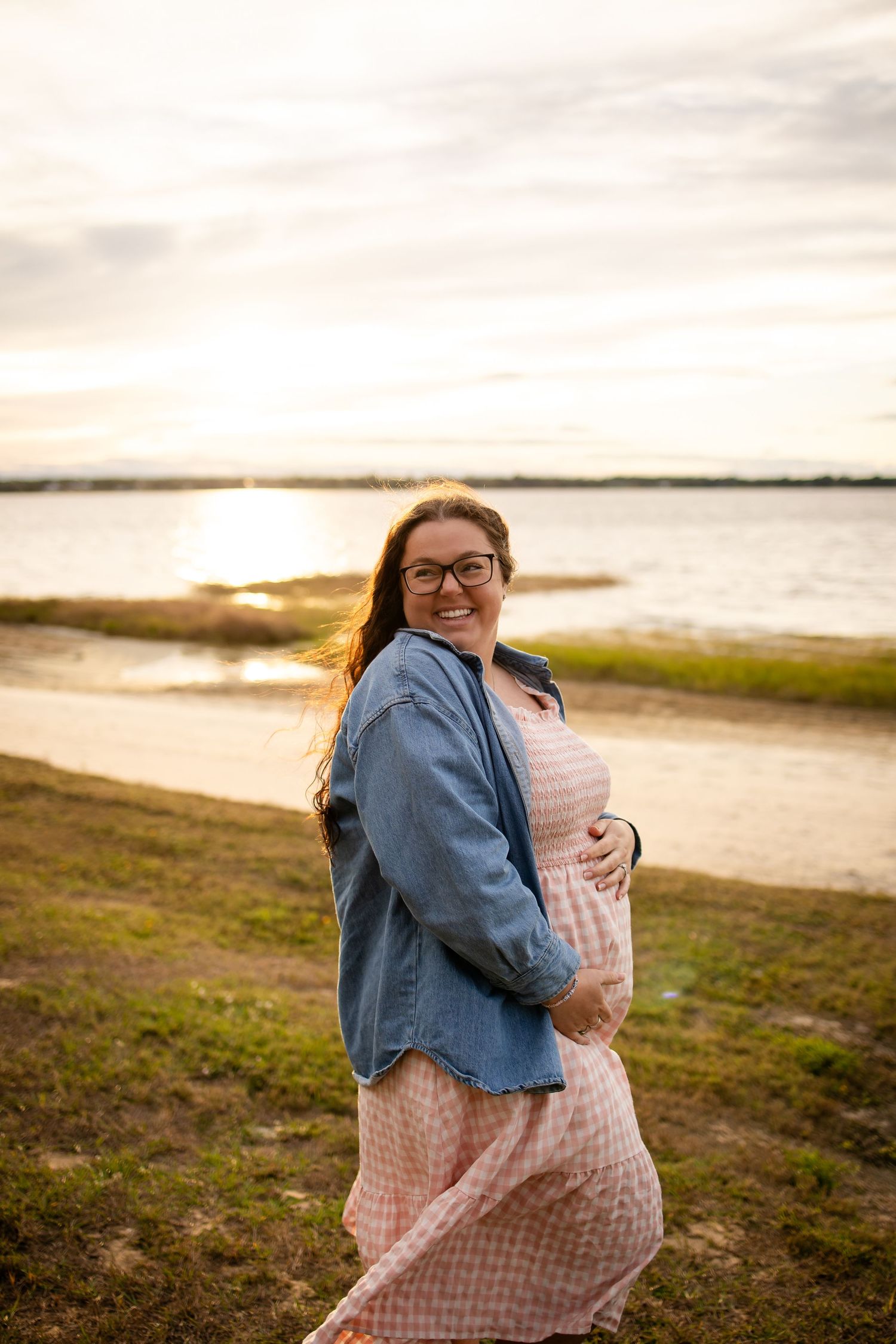 Person in pink gingham dress and denim jacket laughing joyfully by the water at sunset.