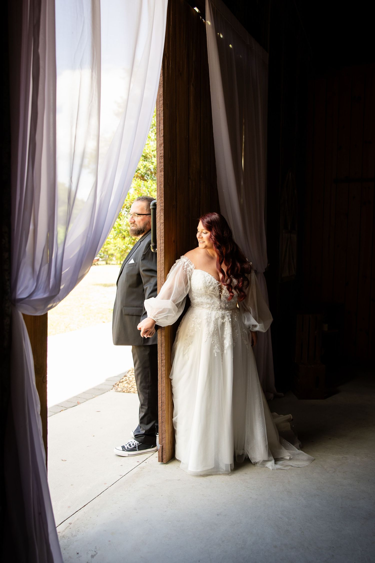 Bride in white lace gown and groom in gray suit hold hands at barn venue doors with sheer curtains.