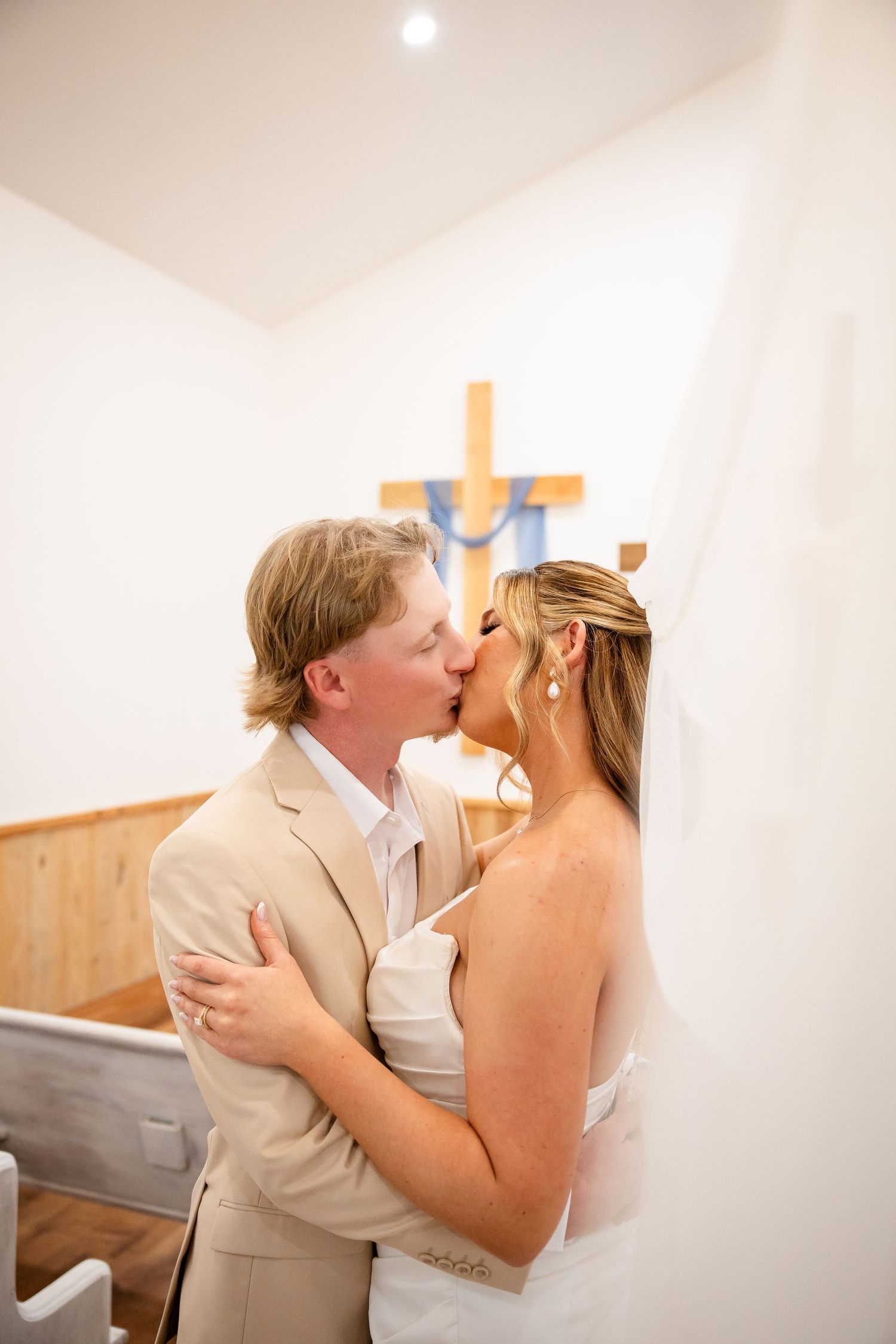 Bride and groom share a kiss in a small white chapel with a wooden cross, bride wearing veil and white dress.