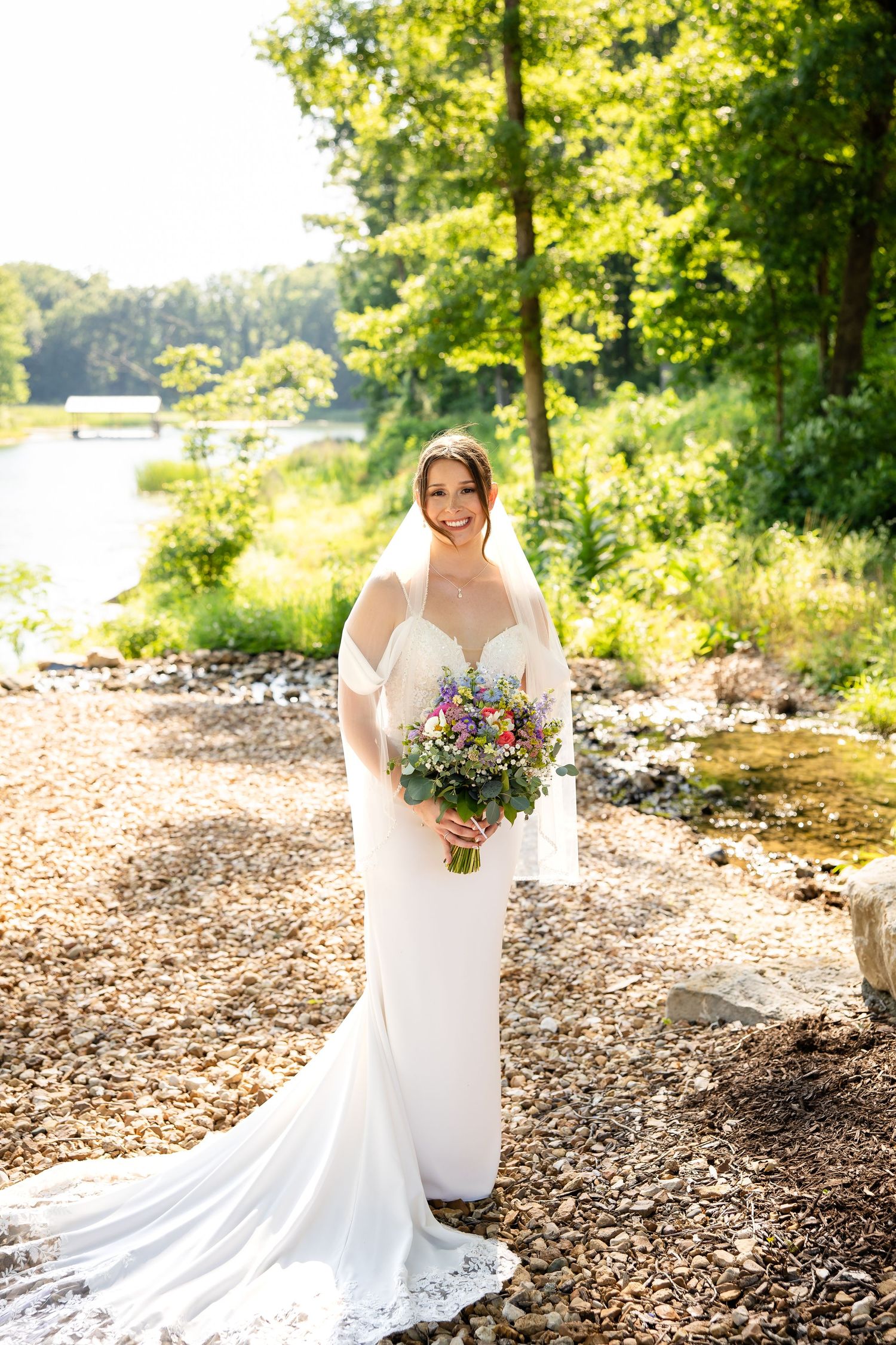 Smiling bride in white gown holding colorful bouquet standing near a sunlit lakeside with lush green trees.