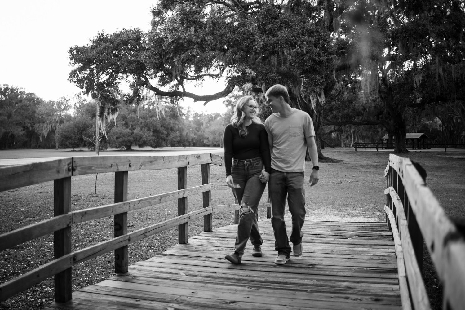 People stroll along a wooden boardwalk surrounded by oak trees.