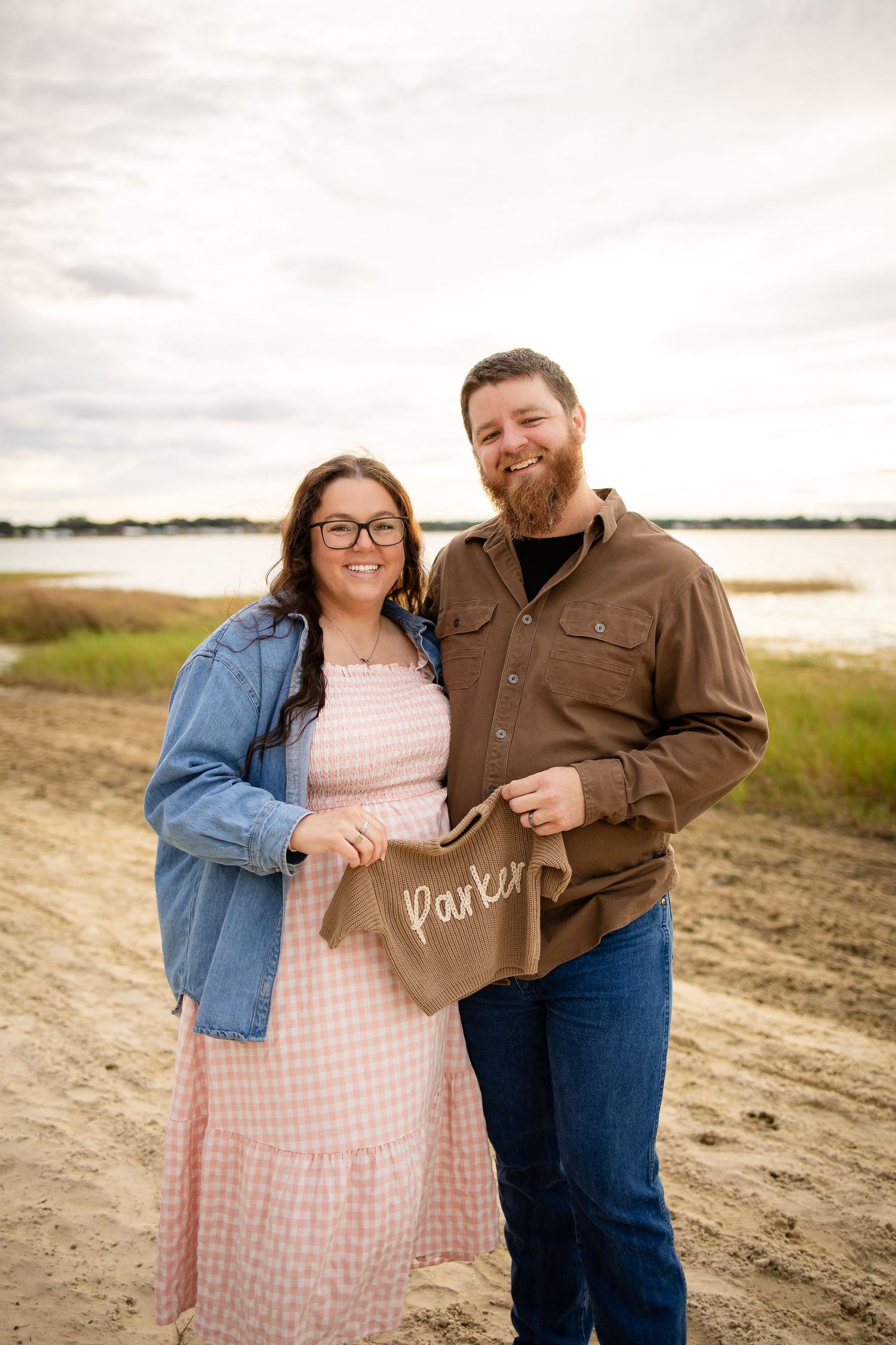 A couple stands on a dirt path by a lake holding a sign that says 'Parents' on an overcast day.