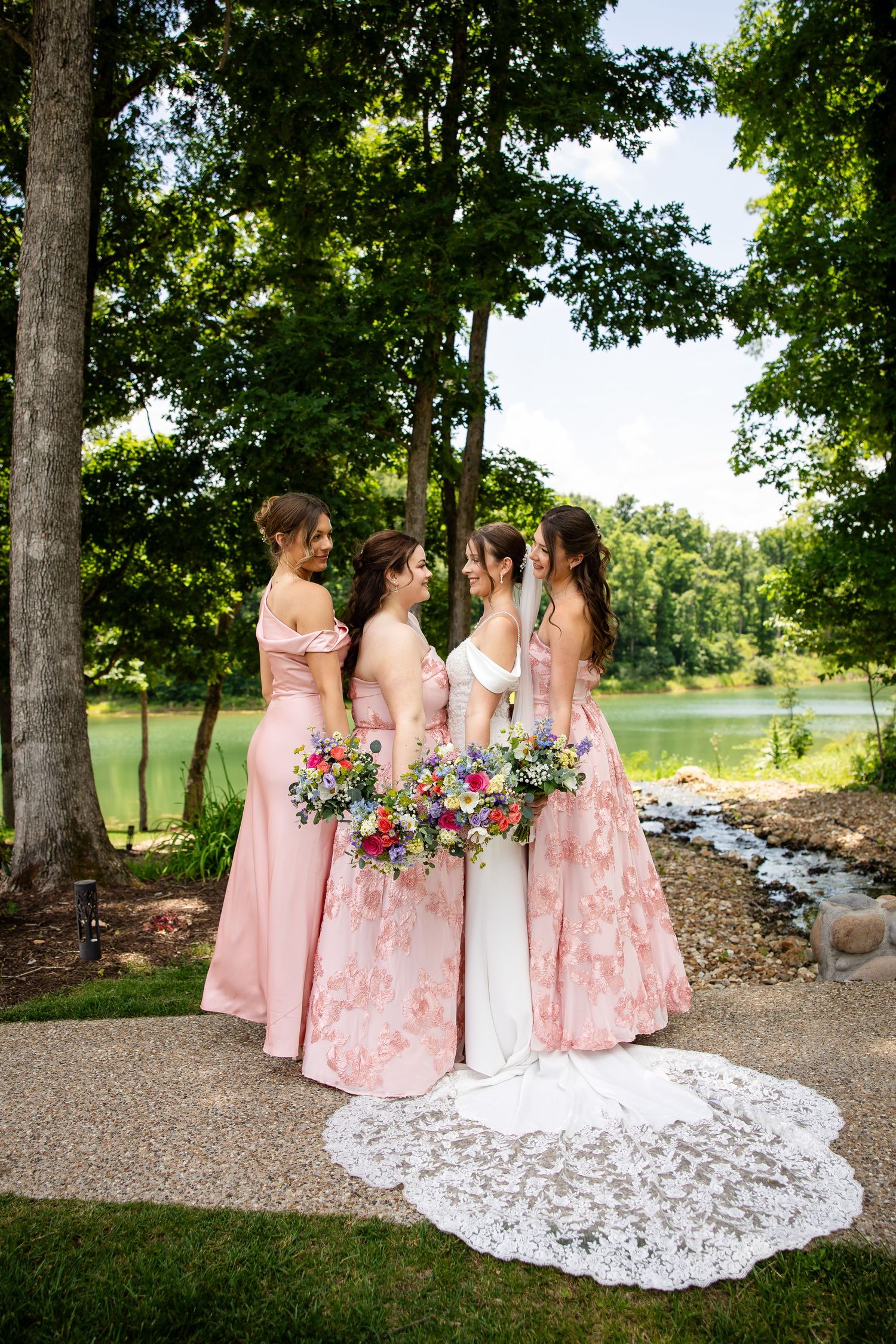 Bride in white gown with lace train stands with three bridesmaids in pink floral dresses holding colorful bouquets outdoors.