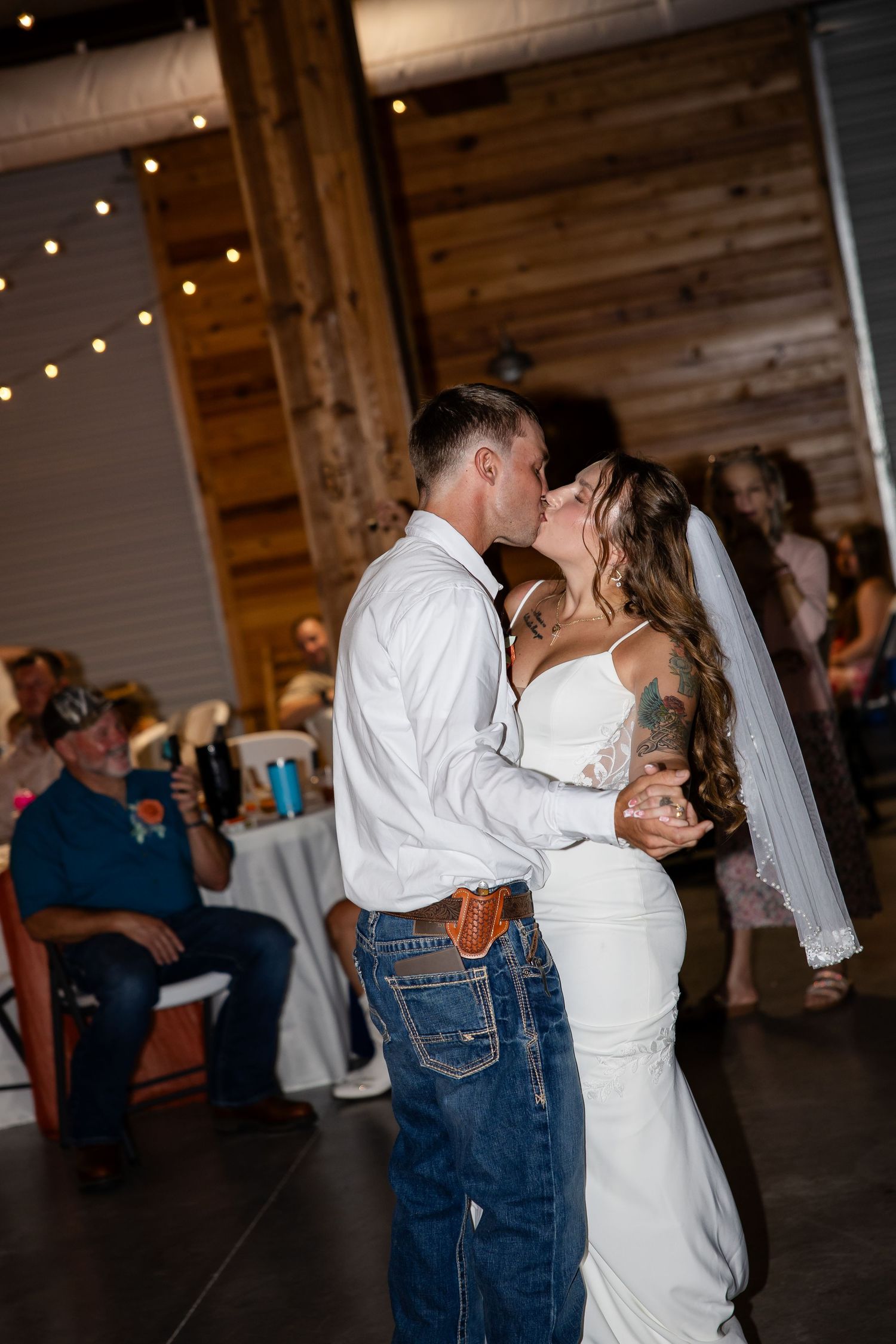 Bride and groom share first dance kiss at rustic barn wedding reception with string lights.