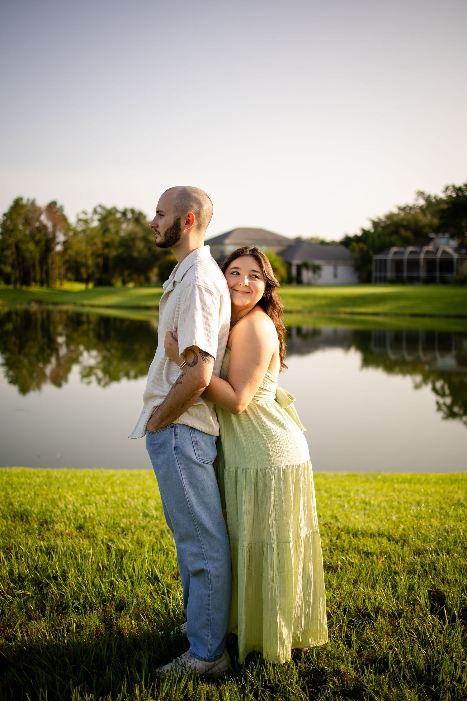 A couple stands back to back near a serene lake at sunset, with the pregnant woman wearing a flowing yellow dress.