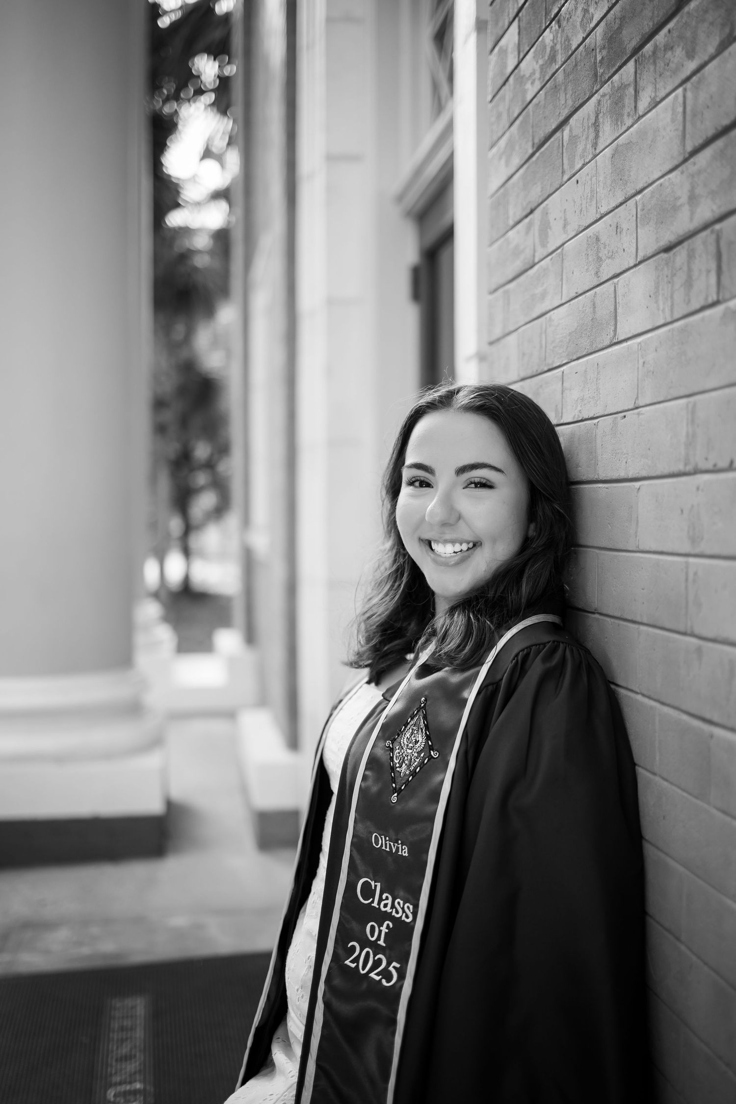 Black and white graduation portrait of a graduate in academic regalia smiling while leaning against a brick wall.