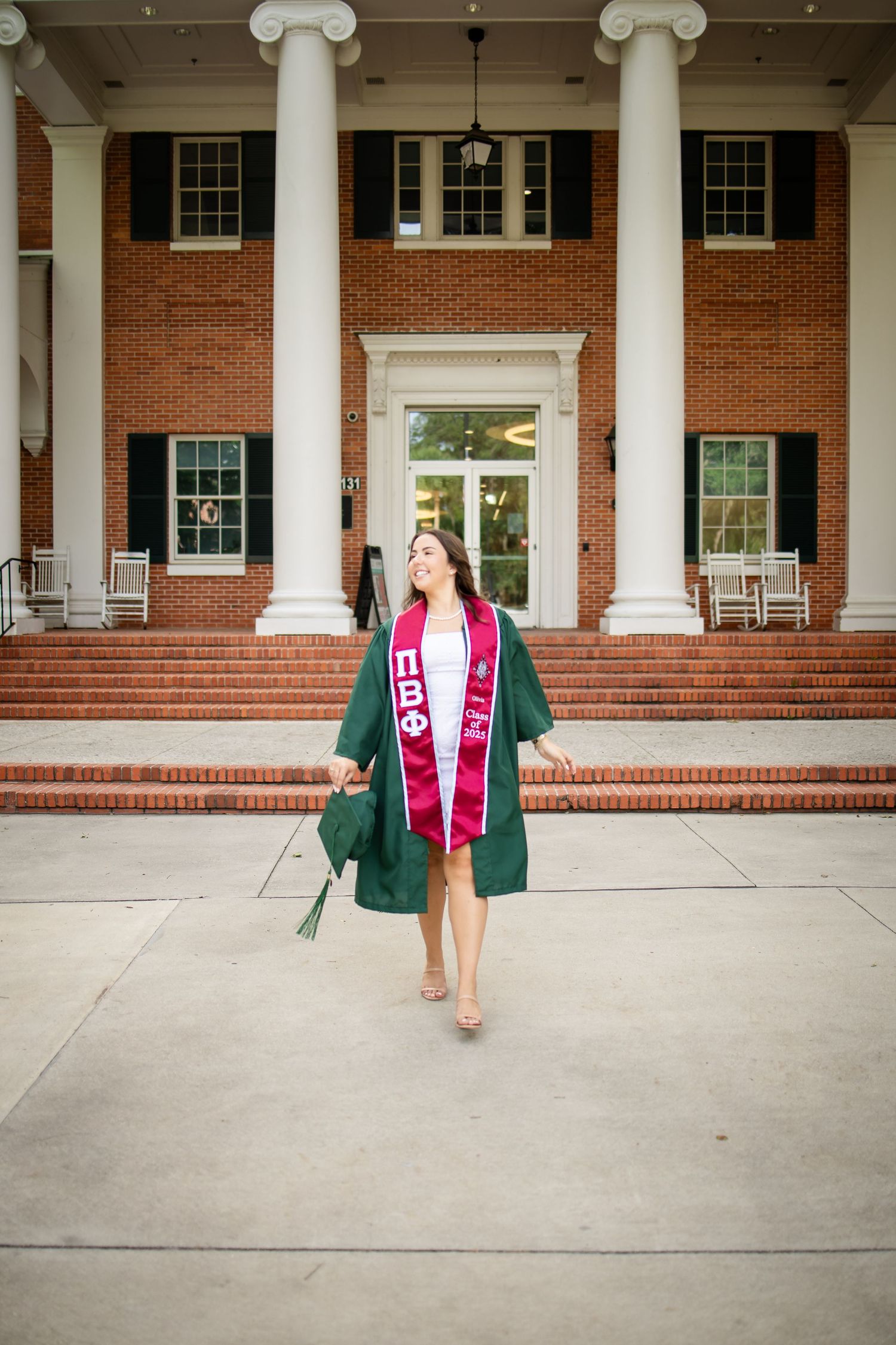 Graduate walking down stairs of columned brick building wearing green cap and gown with pink stole.