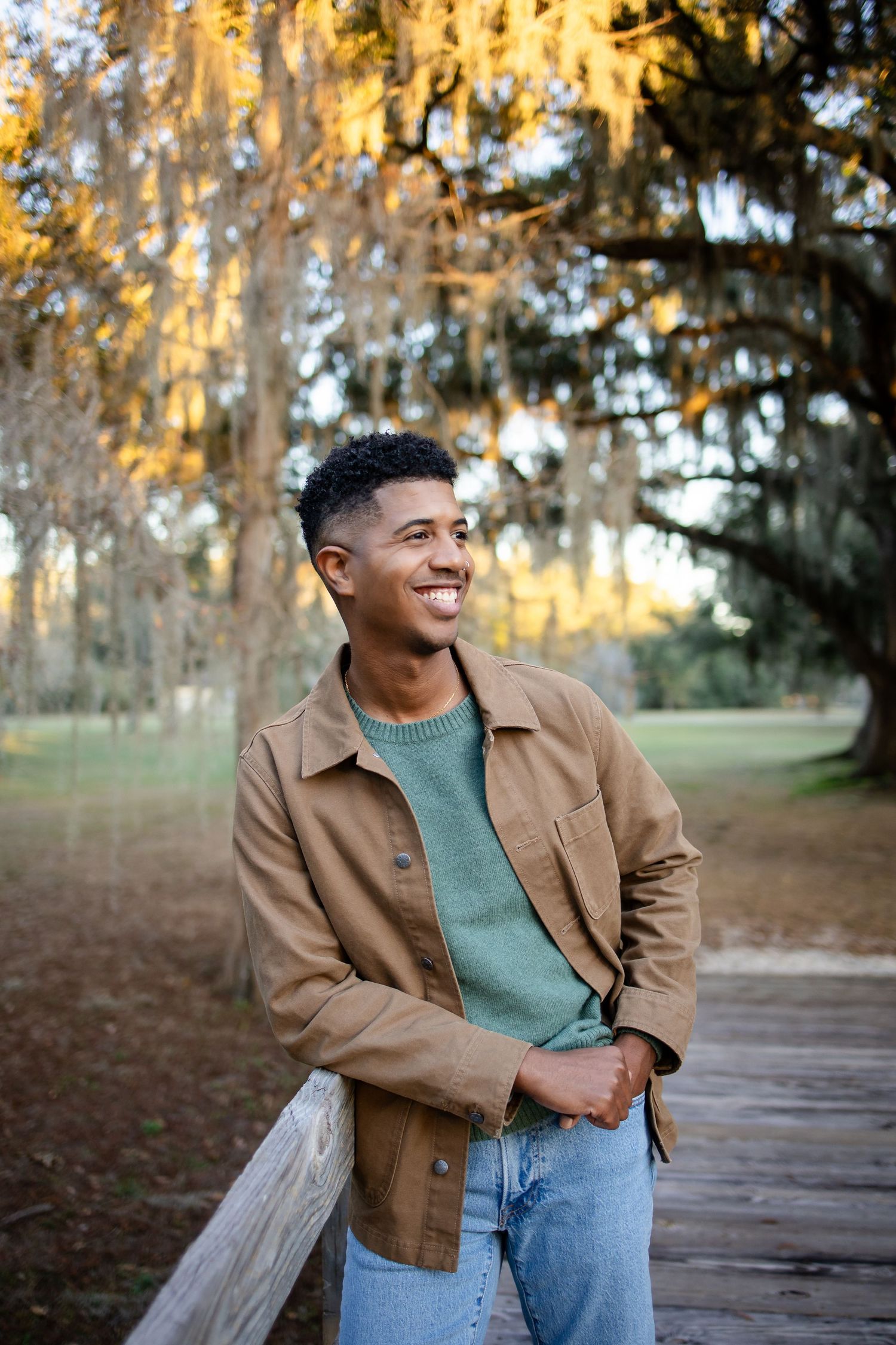Smiling person in tan jacket leans against wooden fence with Spanish moss trees in background.