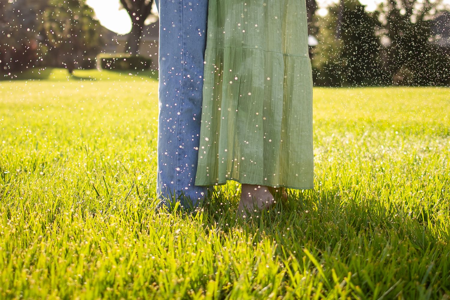 Two people wearing long dresses stand in a sunlit grassy field with sparkling water droplets in the air.