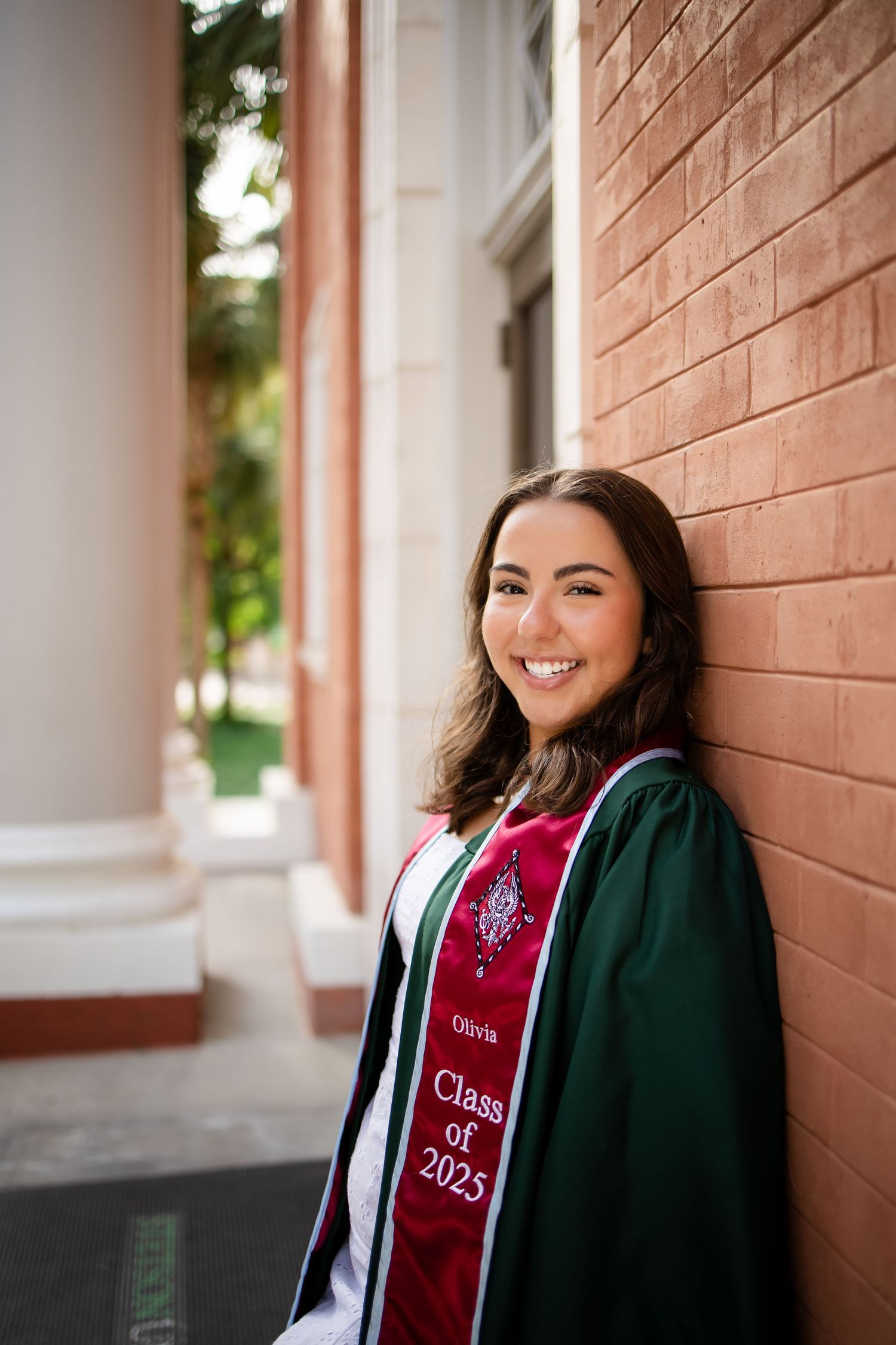 Graduate in green and red academic regalia poses against brick wall during commencement.