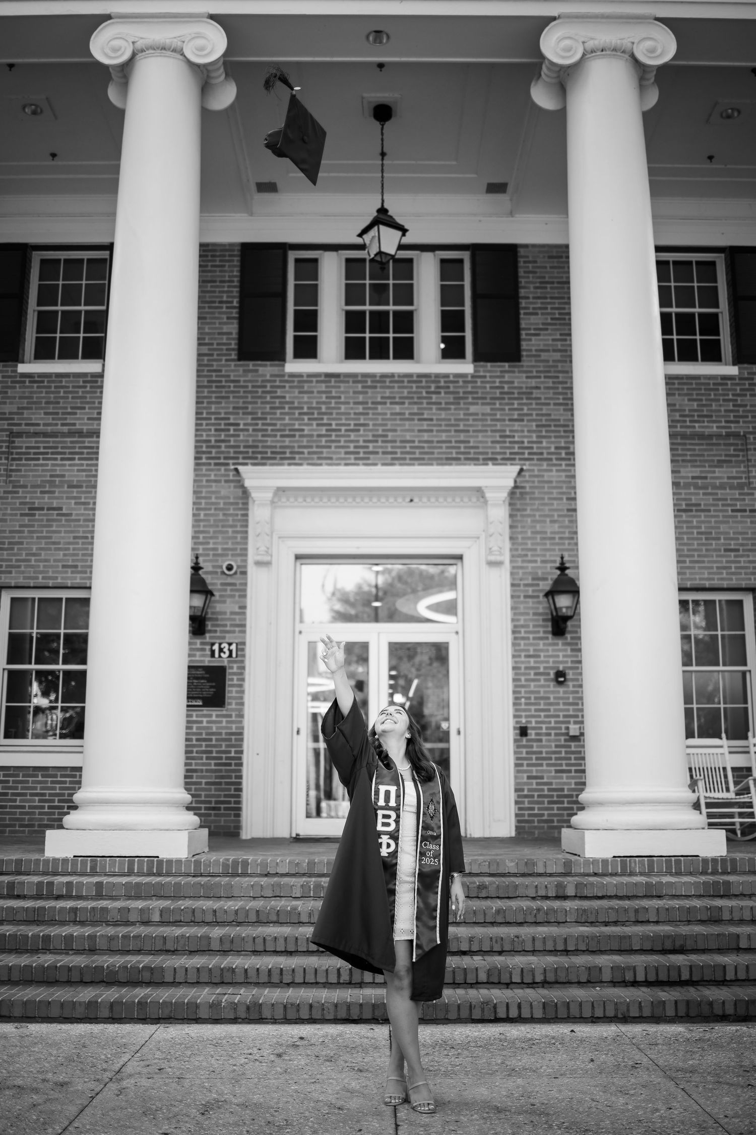 Graduate stands in front of grand brick building with white columns and black shutters on graduation day.