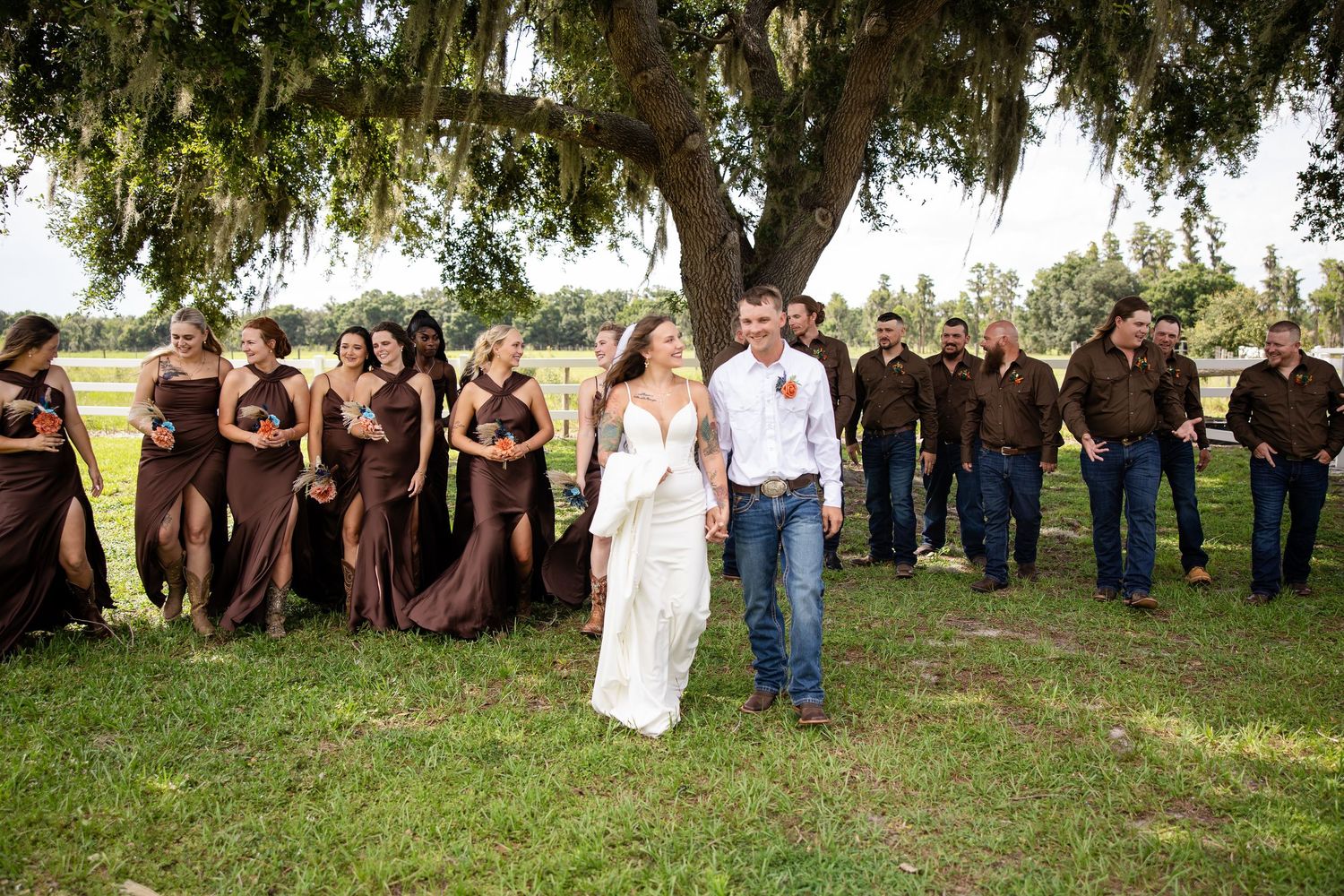 Bride and groom walk together at rustic outdoor wedding with bridesmaids in brown dresses and groomsmen in jeans under oak tree.