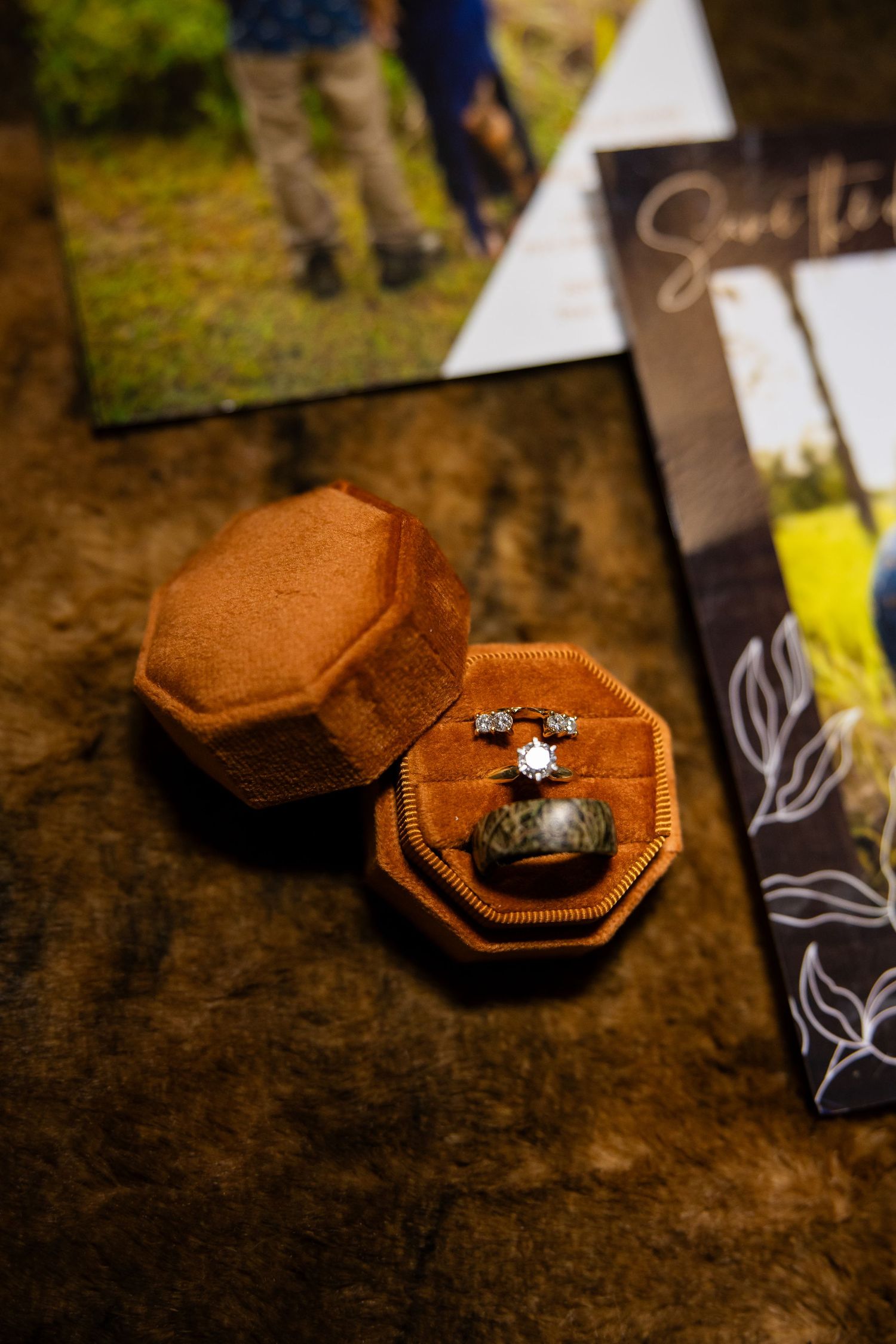 Orange velvet ring box with diamond engagement ring, earrings, and camo band beside save the date cards on wood.