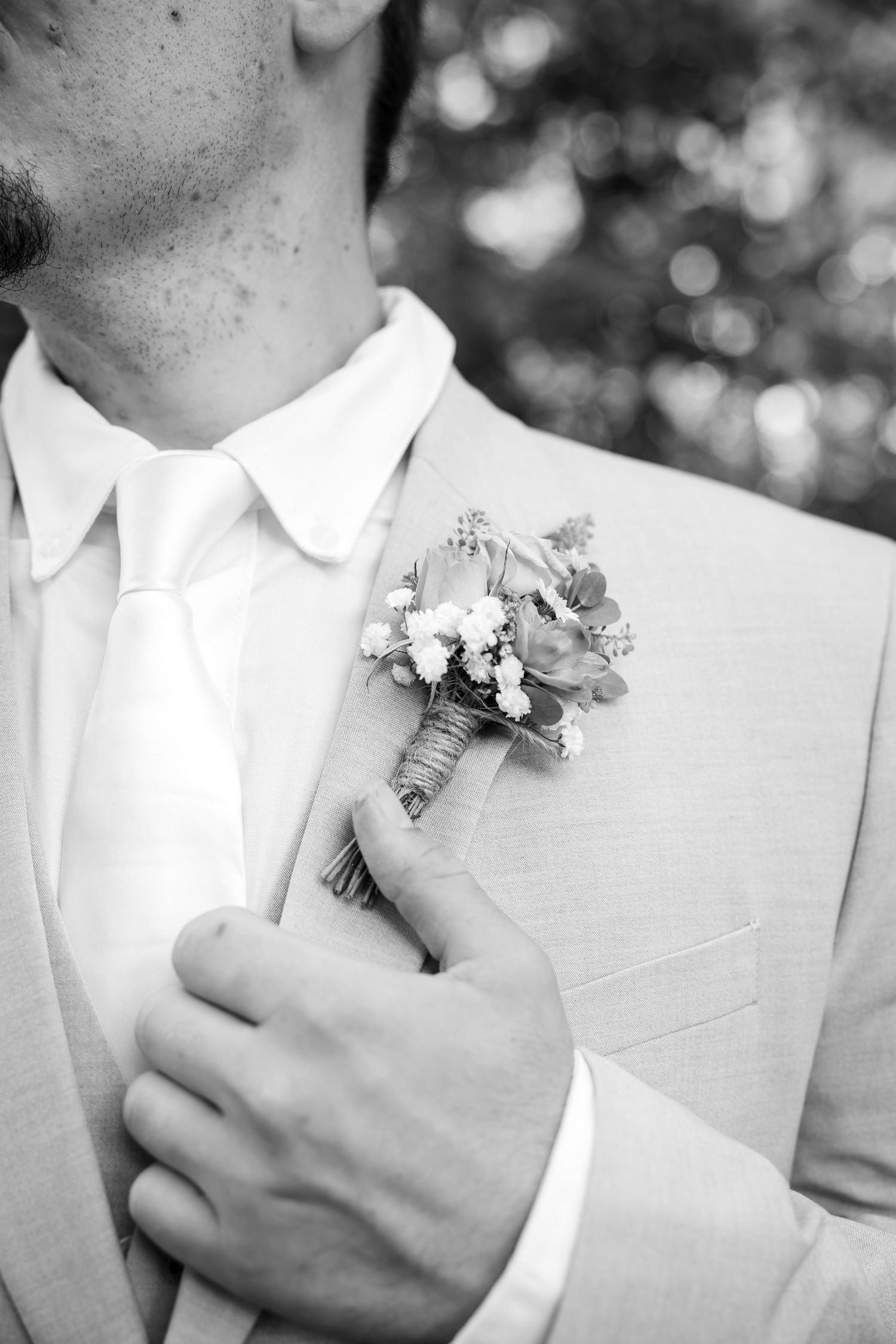 Black and white photo of a groom in a light suit adjusting a floral boutonniere on his lapel.
