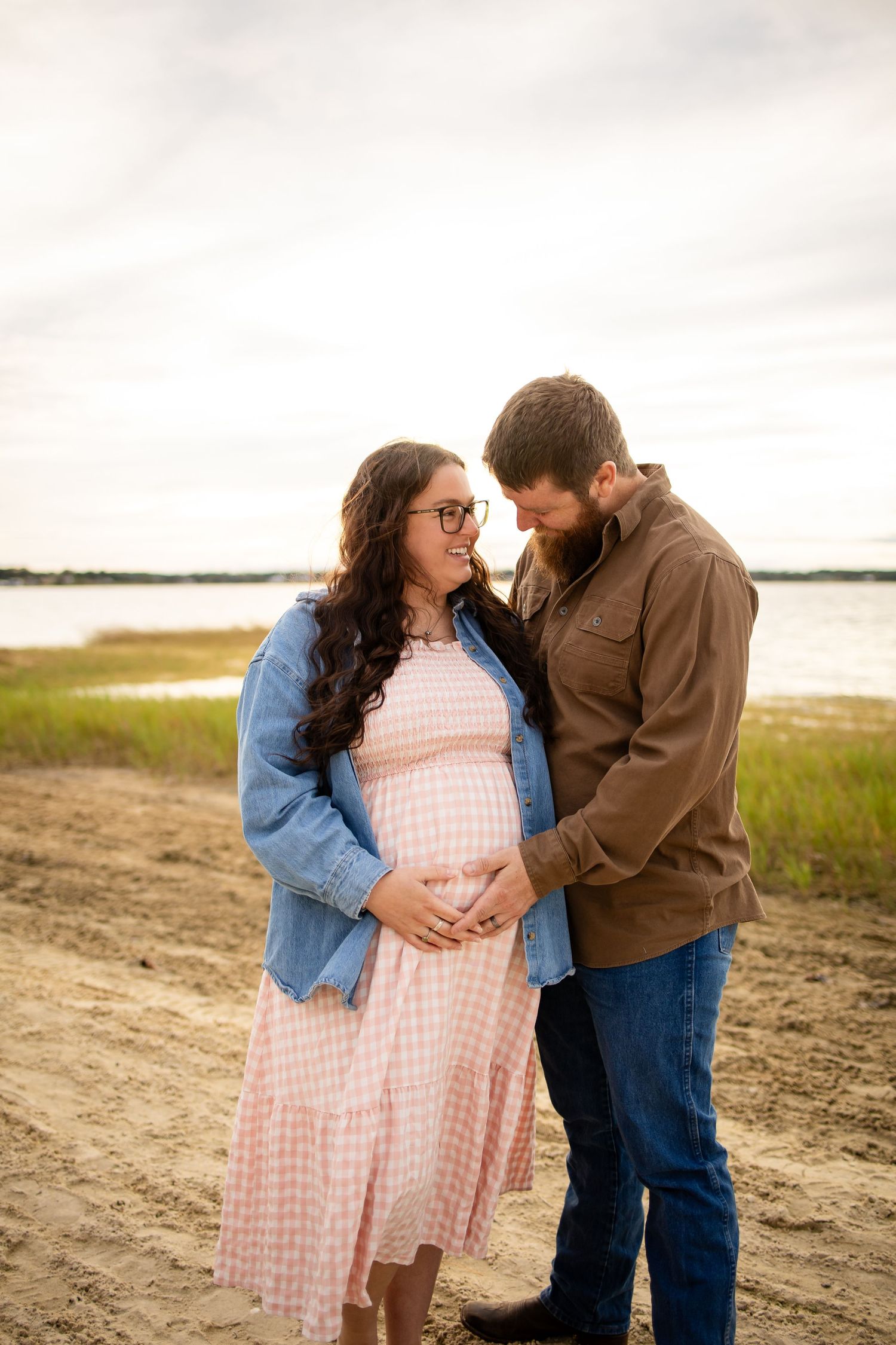 A couple shares an intimate moment on a beach during a maternity photoshoot at sunset.