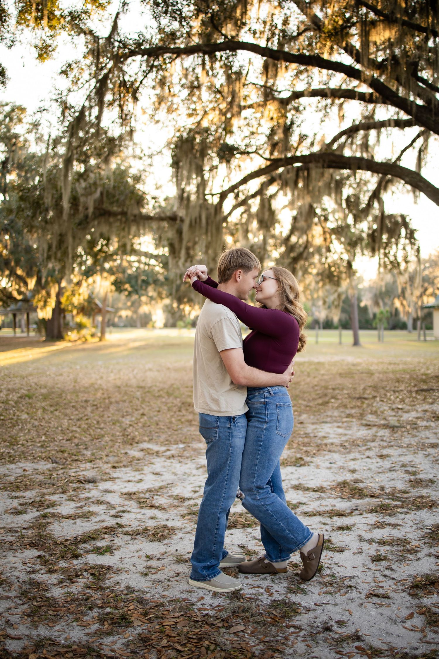A romantic couple embraces playfully in a park with Spanish moss hanging from trees at sunset.