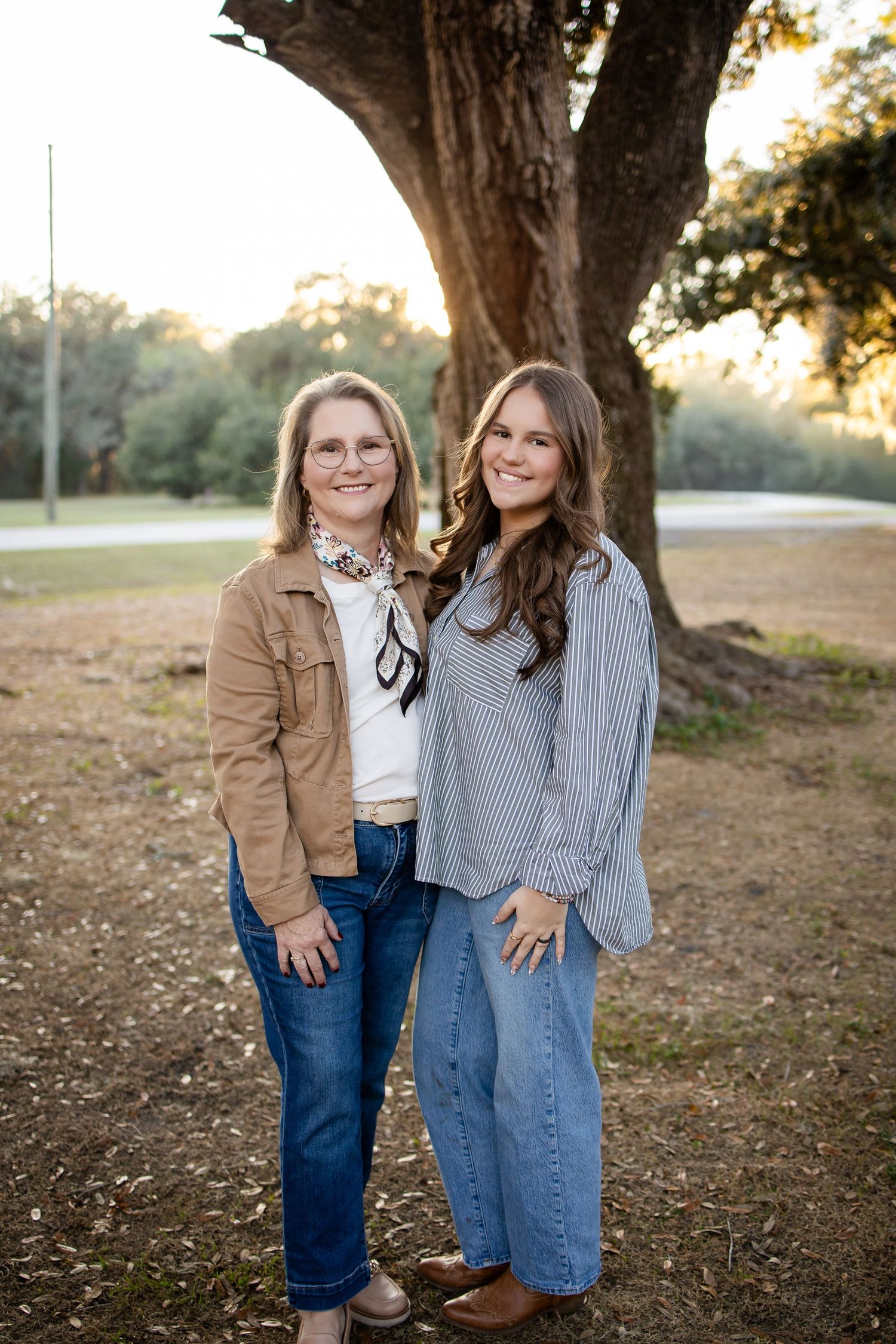 Two people in coordinated casual outfits share joyful moments under large oak trees at sunset.