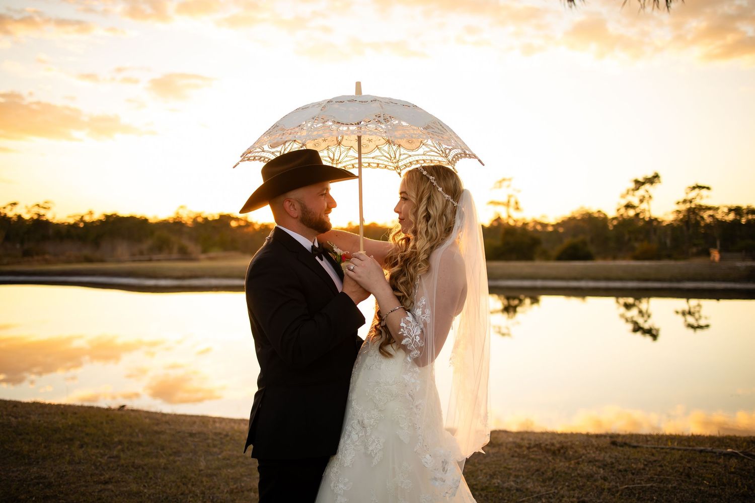Bride and groom share a romantic sunset moment under a lace umbrella near a reflective pond at their outdoor wedding.