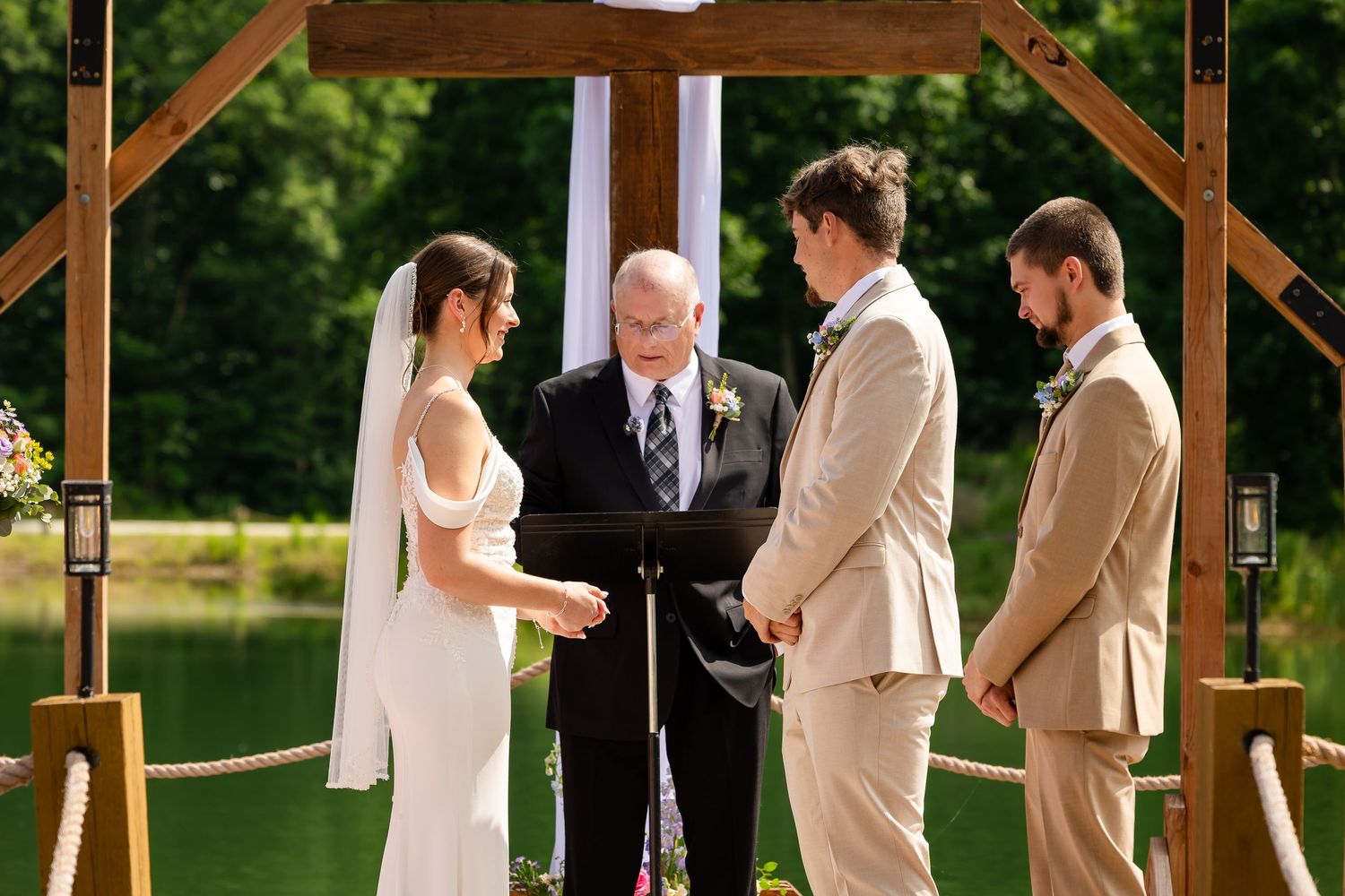 Outdoor wedding ceremony by a lake with bride, groom, best man, and officiant under a wooden arch with white draping.