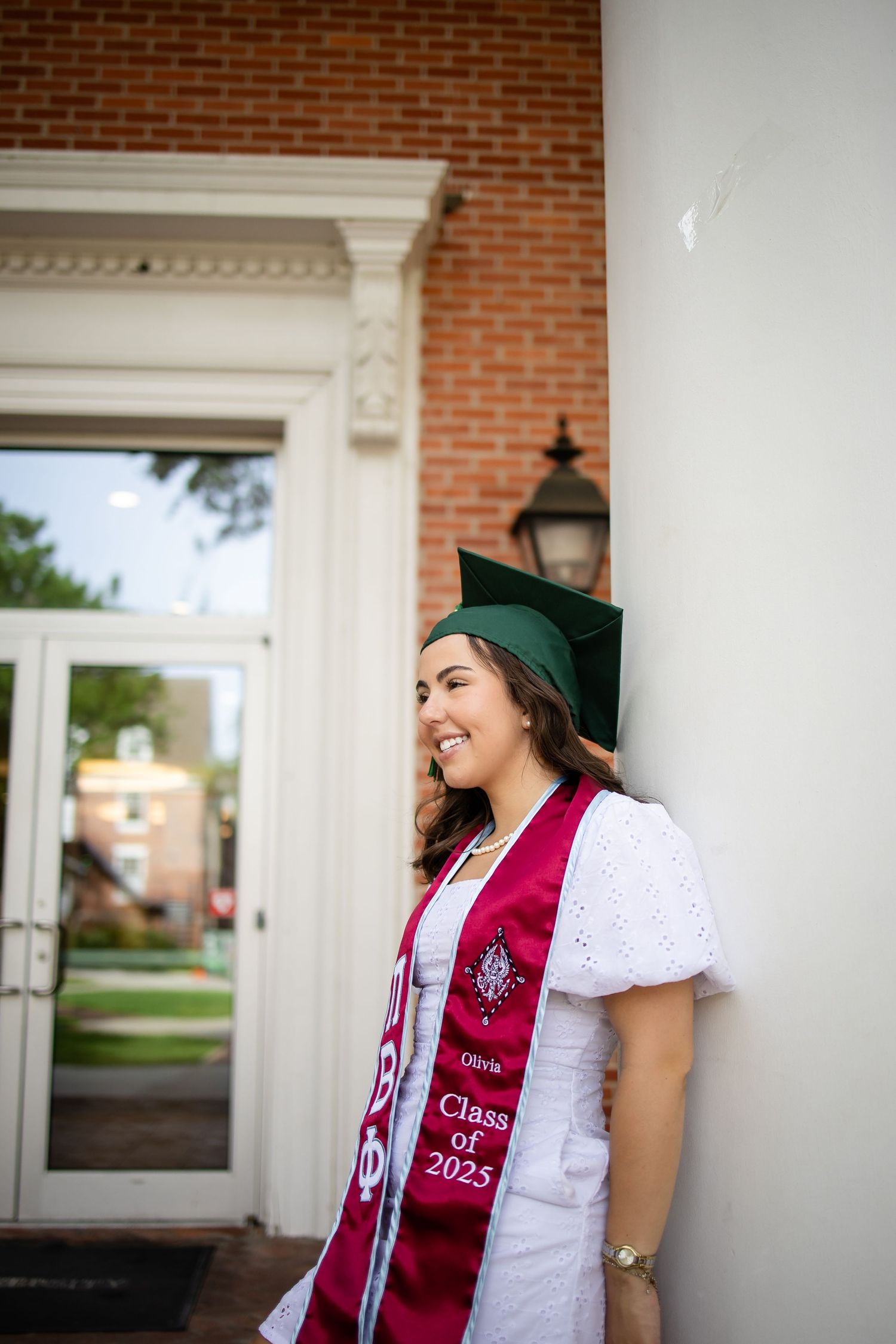 A graduate in cap and white dress stands smiling by a brick building entrance wearing a graduation stole.