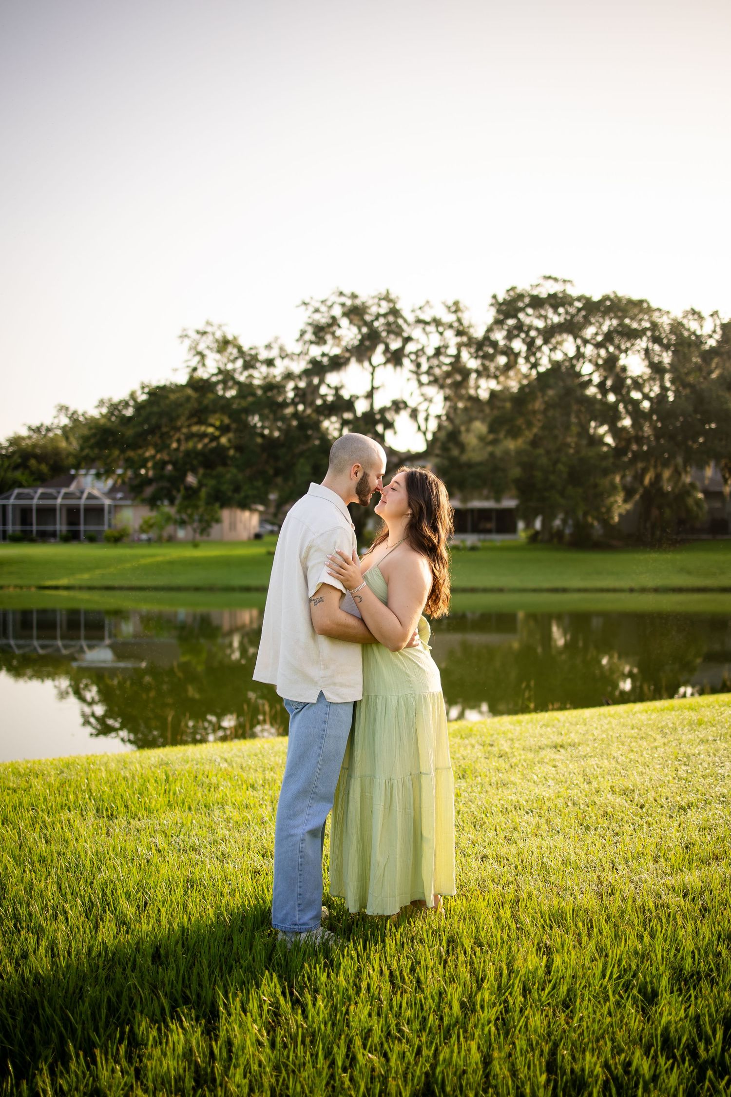 Romantic outdoor scene at sunset with a couple embracing on a grassy hill beside a pond.