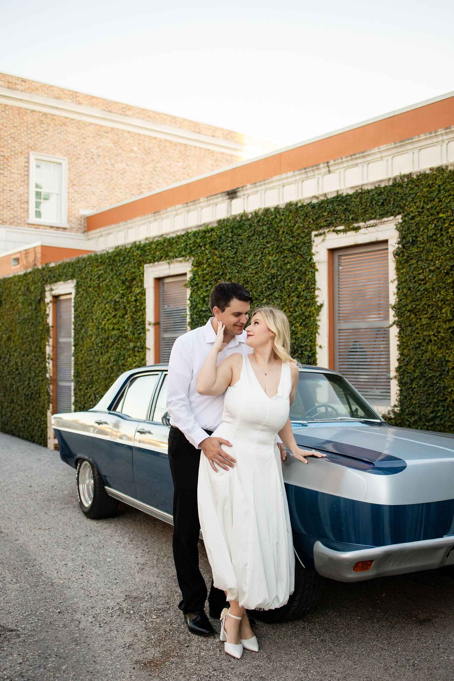 A couple in wedding attire pose together against a vintage car parked by an ivy-covered brick building.