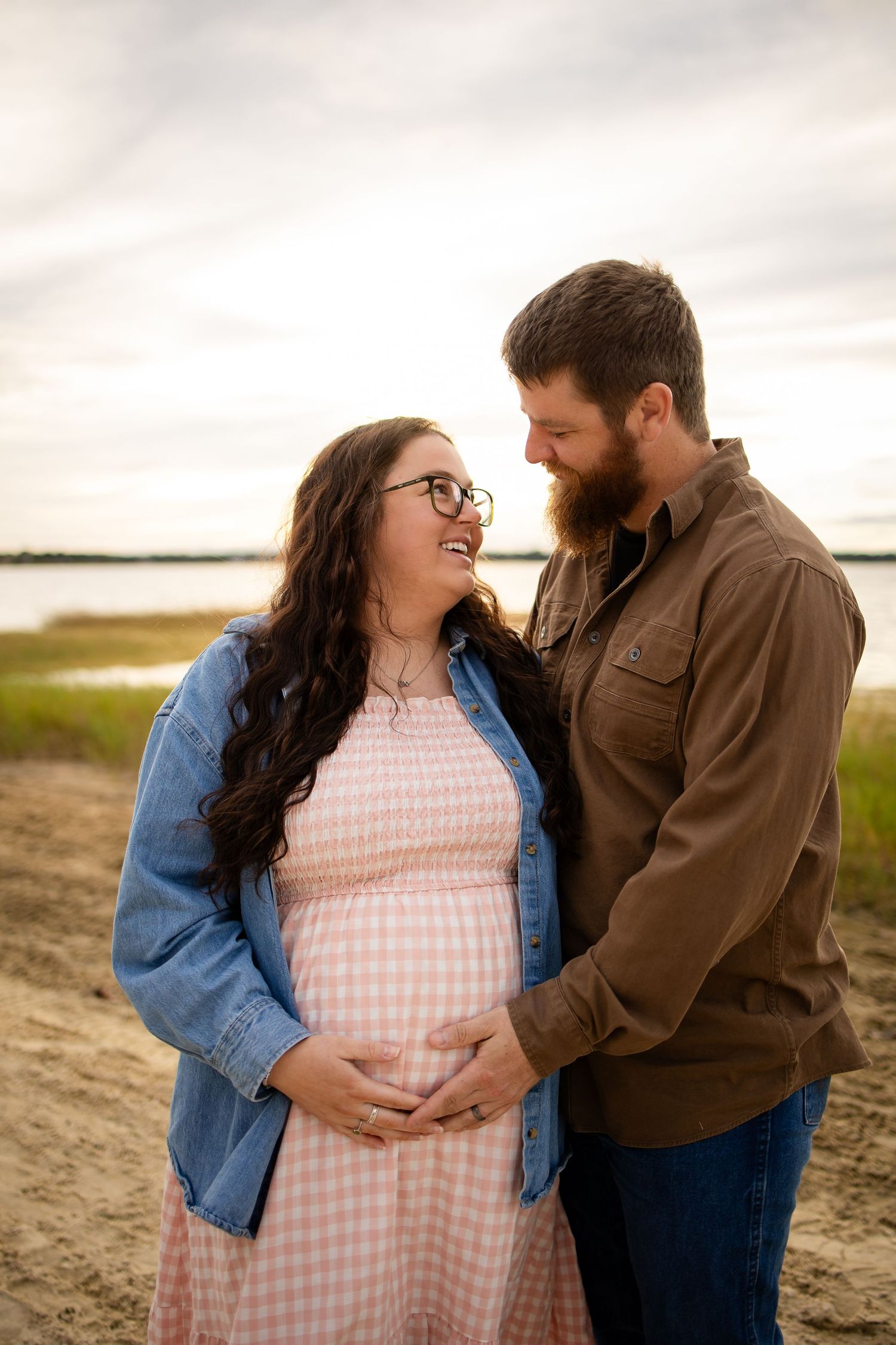 A couple shares an intimate moment while standing near a lake, with the woman wearing a pink gingham dress.