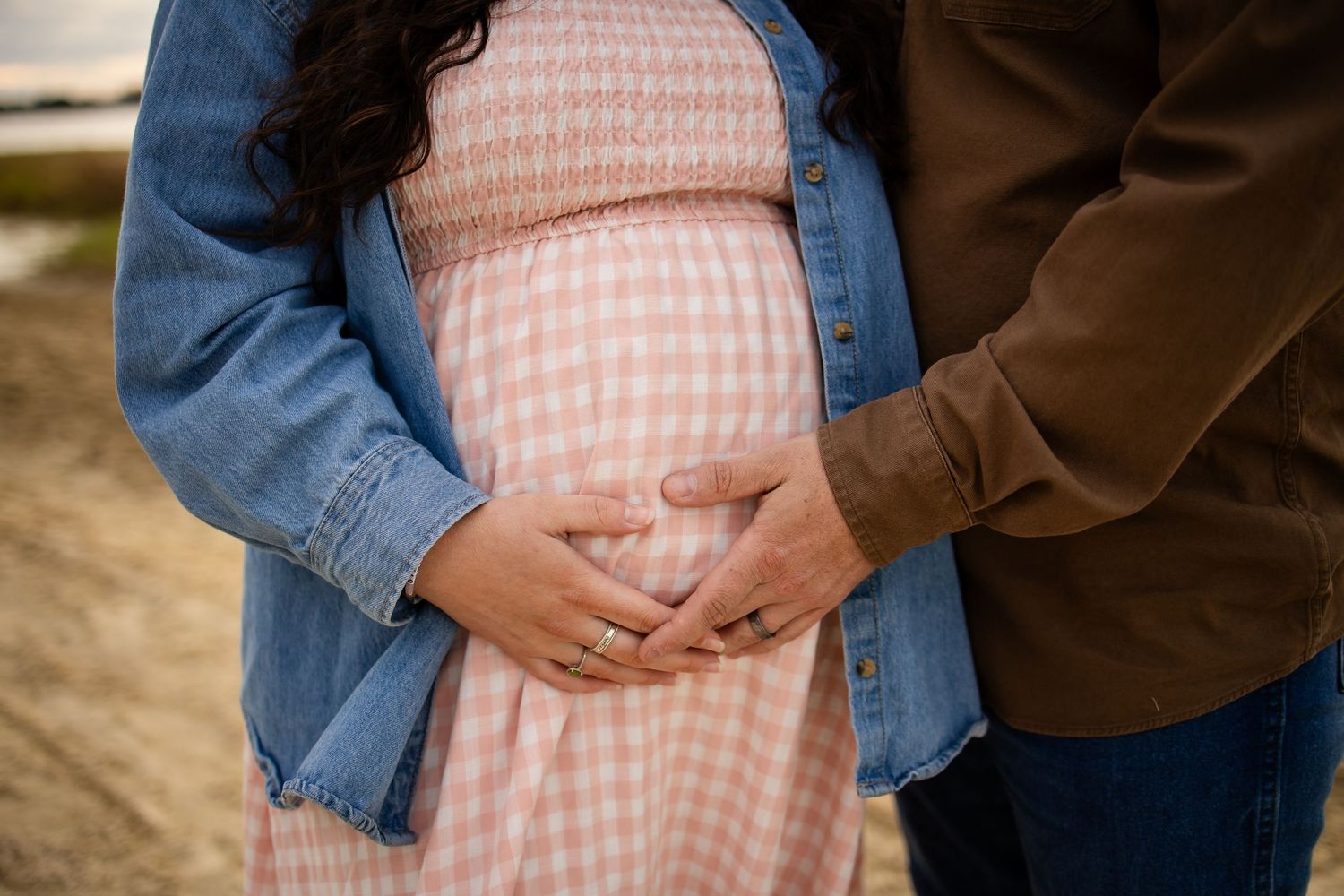 A couple embraces in front of a pink and white gingham dress against a natural outdoor background.