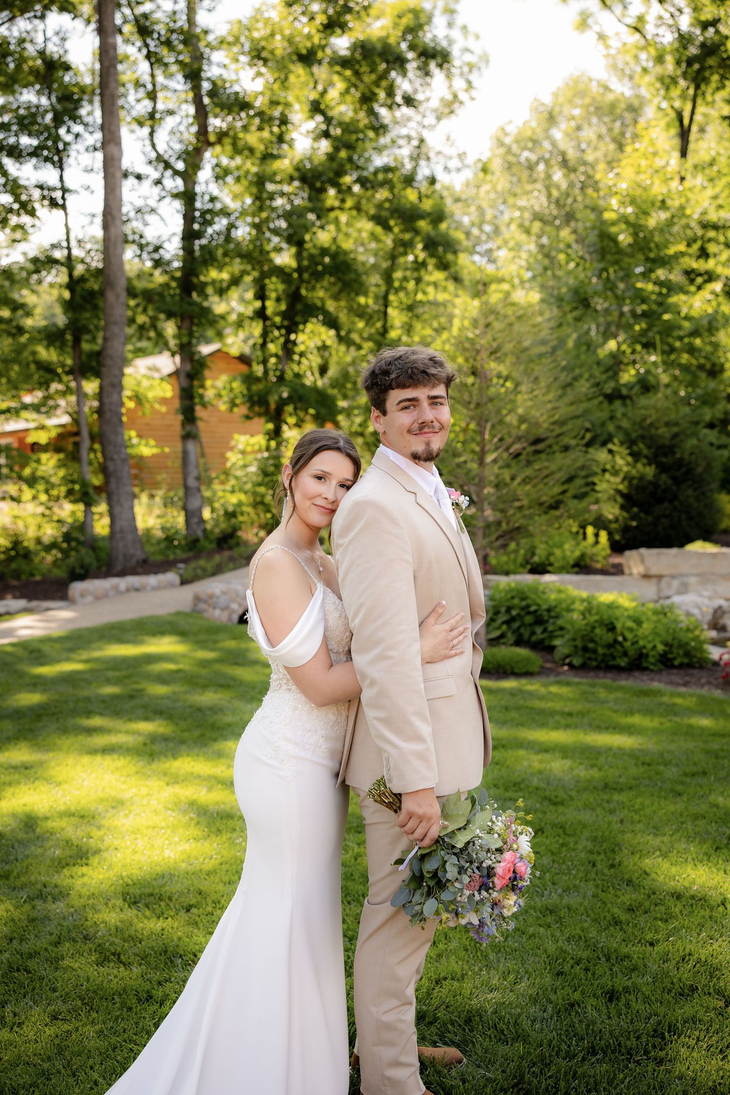 Bride in white off-shoulder gown and groom in beige suit pose back-to-back holding bouquet in lush green garden.
