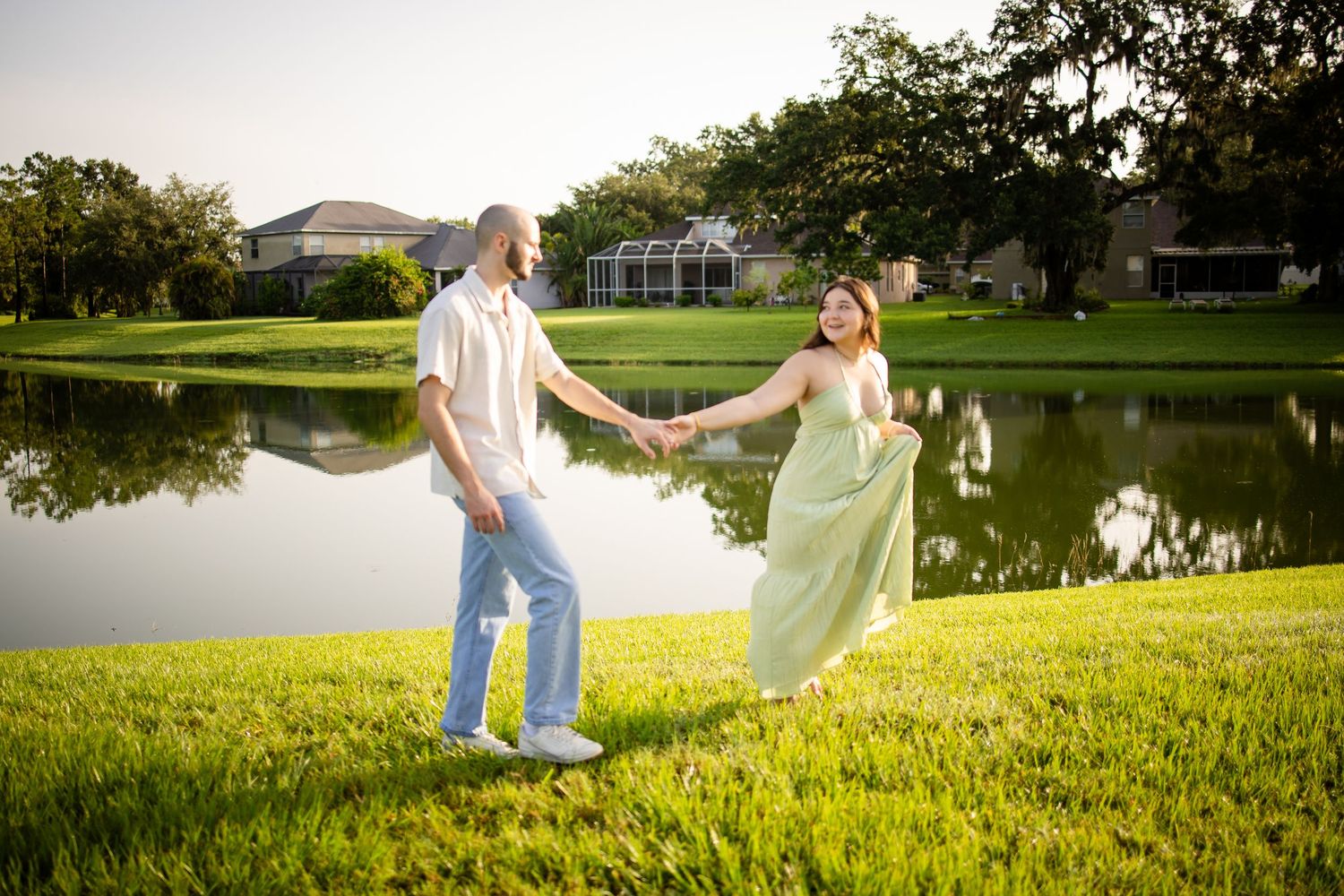 A romantic couple holds hands while dancing on grass near a tranquil pond at sunset.