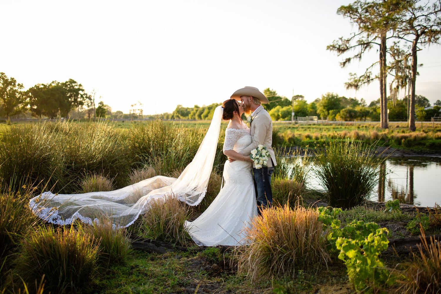 A romantic outdoor sunset wedding photo shoot by a lake with soft golden lighting highlights a couple in a natural setting.