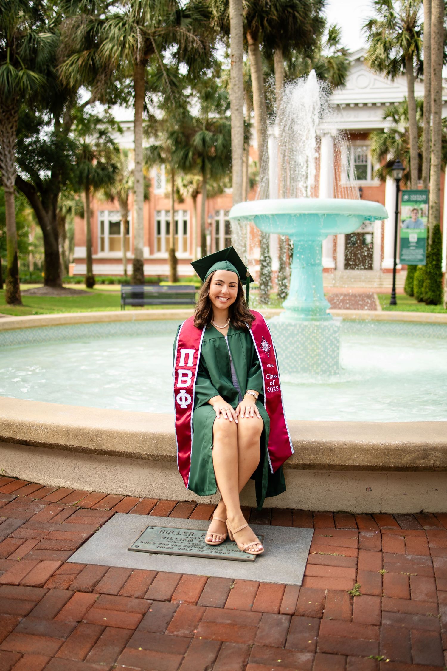 Graduate photos at a university fountain featuring someone in academic regalia and a burgundy graduation stole on brick pavers.