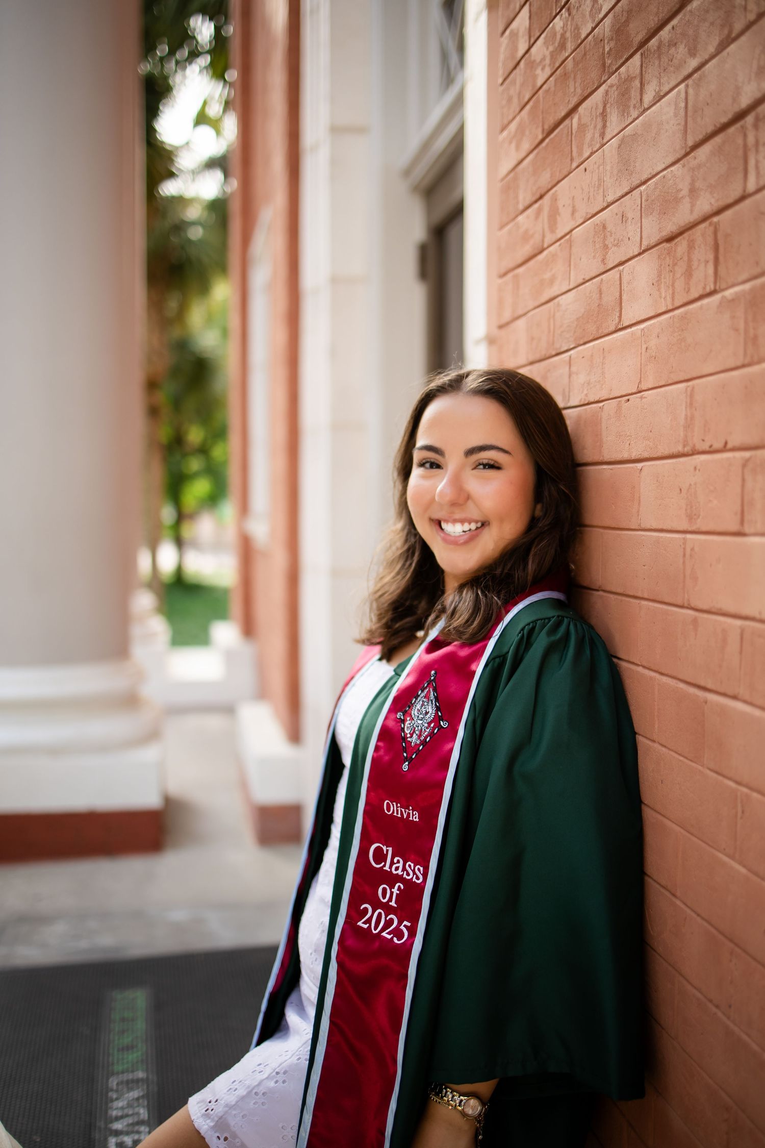 Graduate in green cap and gown with red graduation sash smiles while leaning against brick wall.