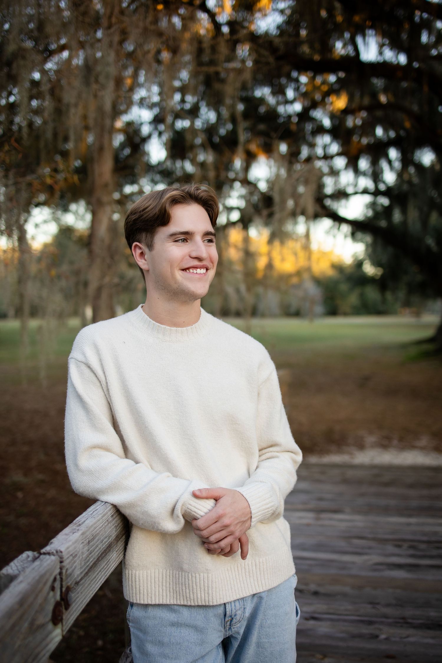 Person in cream sweater smiles while standing on wooden boardwalk with mossy trees in background.