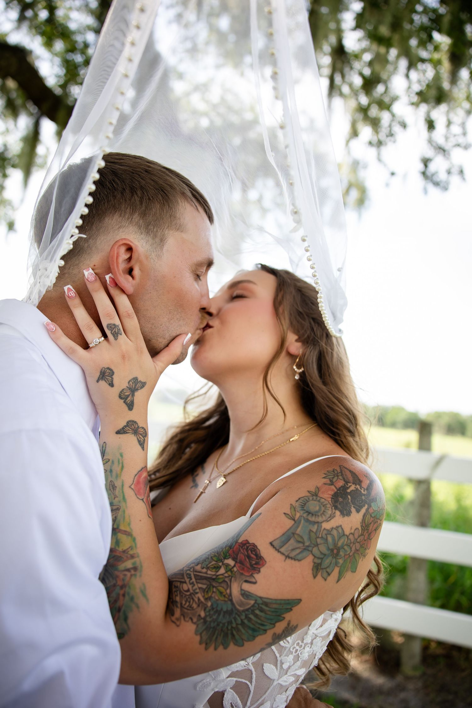 Tattooed bride and groom share a kiss under a white veil outdoors near a white fence.