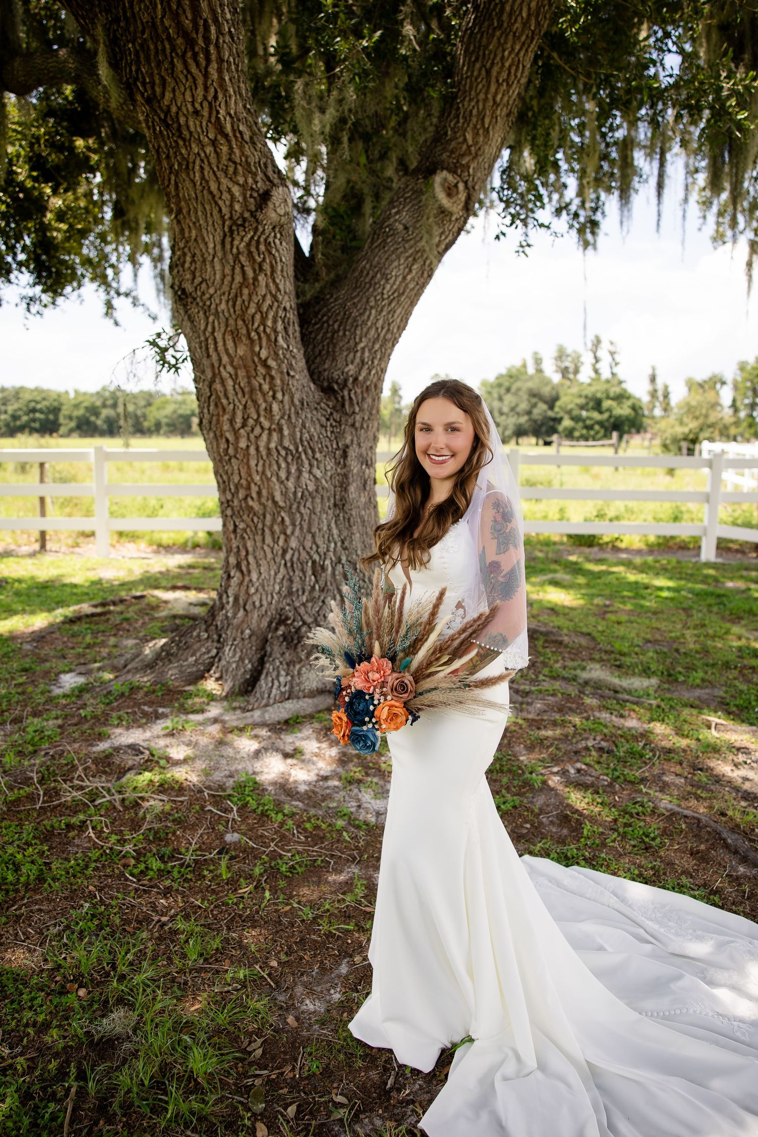 Bride in white gown with veil holds boho bouquet, standing under a large oak tree at an outdoor venue.
