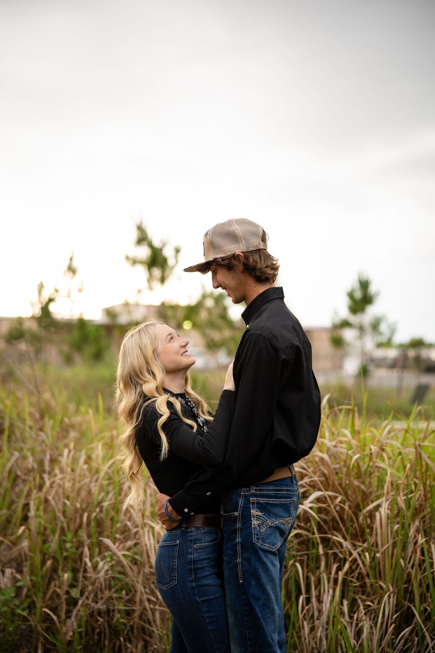 A young couple in jeans and black jackets share a romantic moment in a field of tall grass.