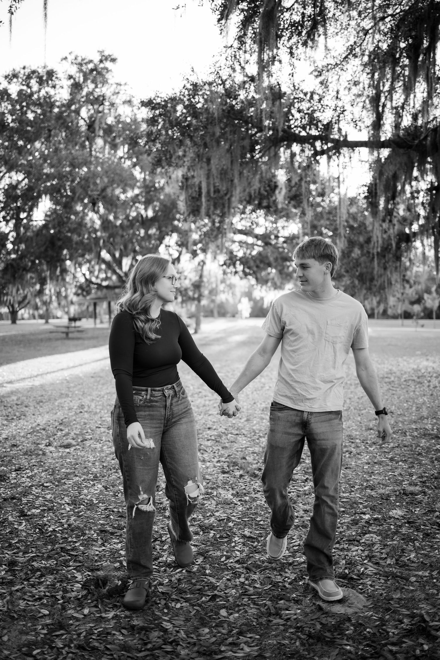 A romantic couple holds hands while walking through a park with Spanish moss hanging from trees in black and white.