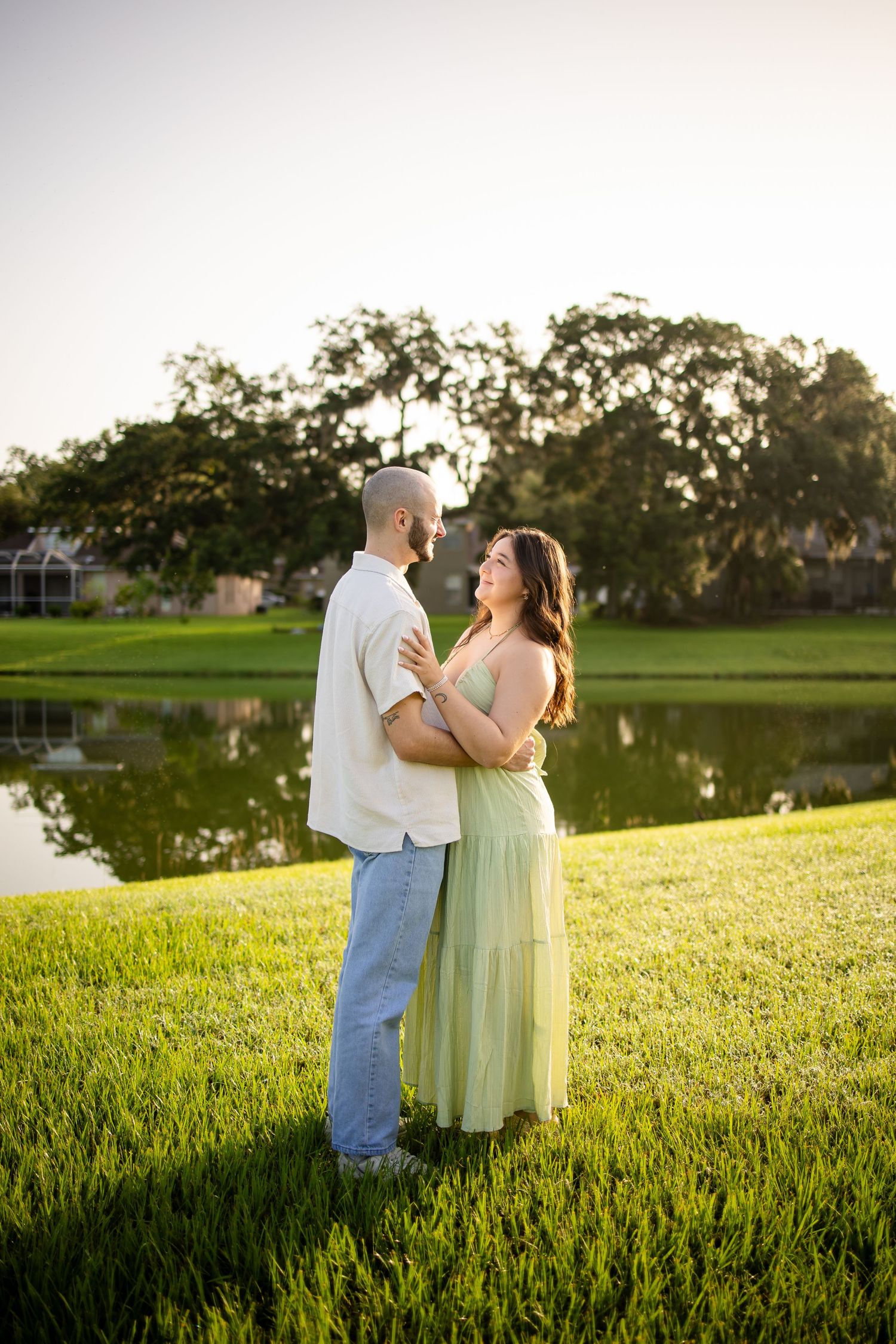 A couple embraces on a grassy hill beside a tranquil pond during golden hour, creating a romantic outdoor scene.