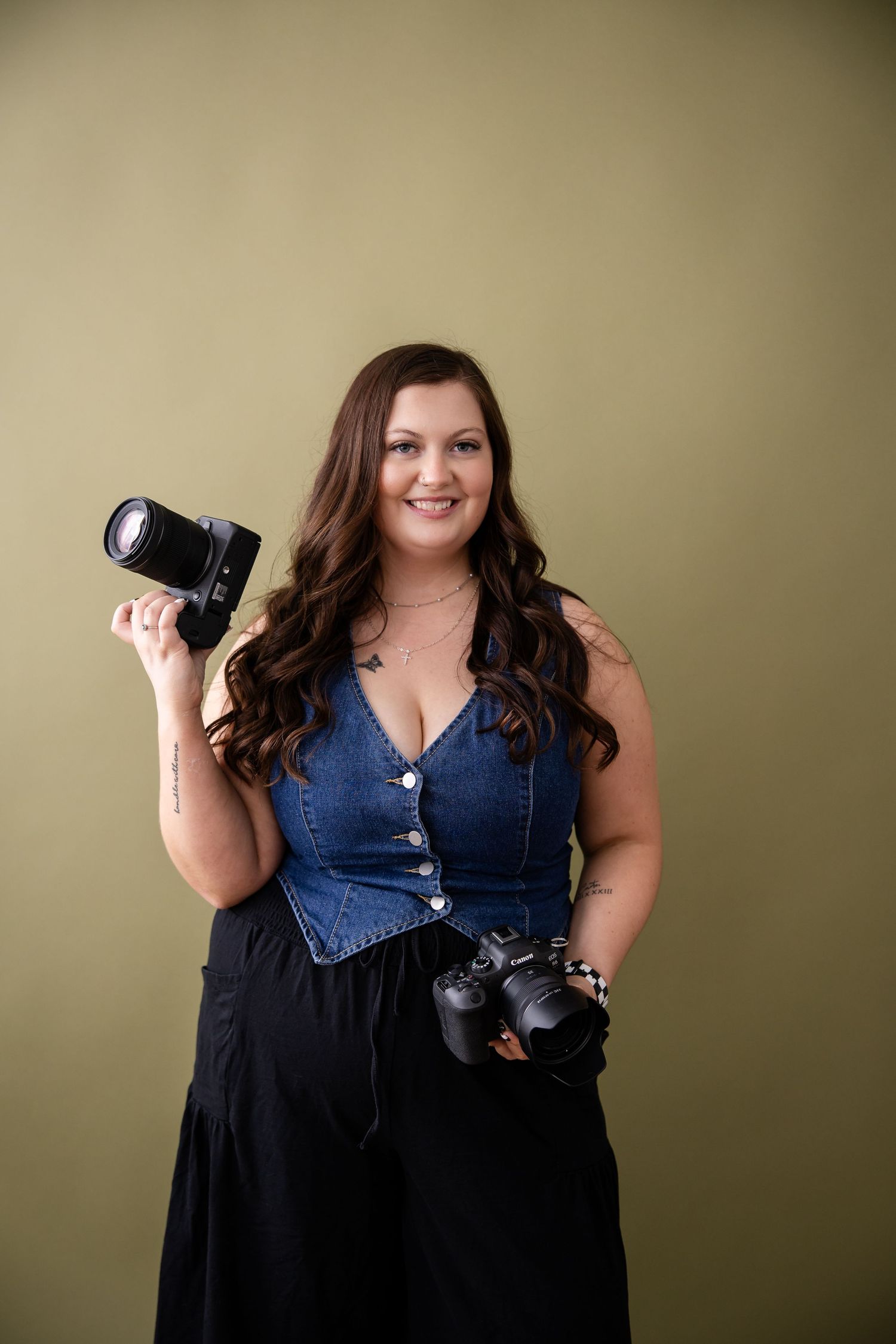 Professional photographer in denim and black outfit poses with camera equipment against neutral background.