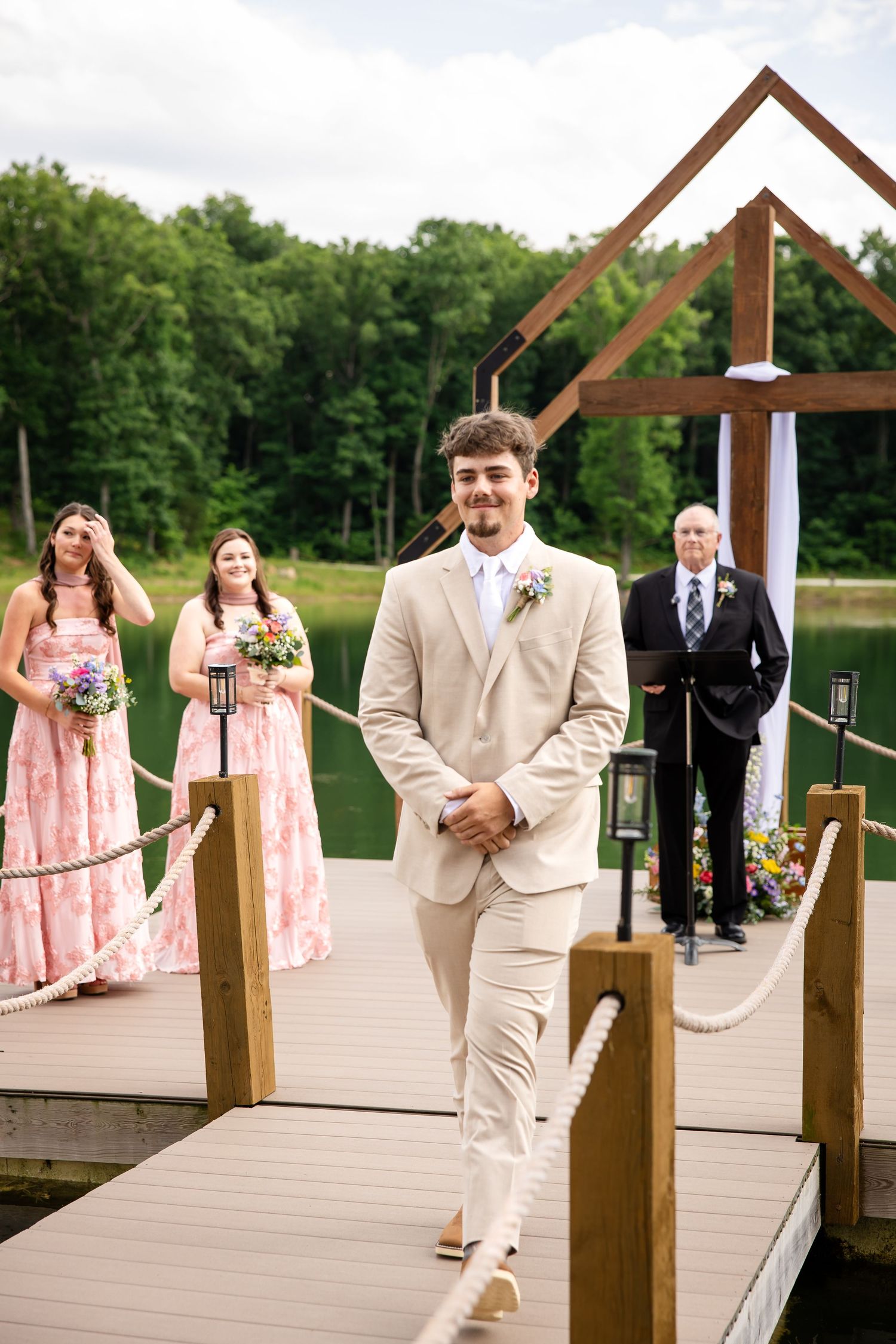 Groom in beige suit walks down dock aisle at outdoor lakeside wedding ceremony with bridesmaids in pink dresses.