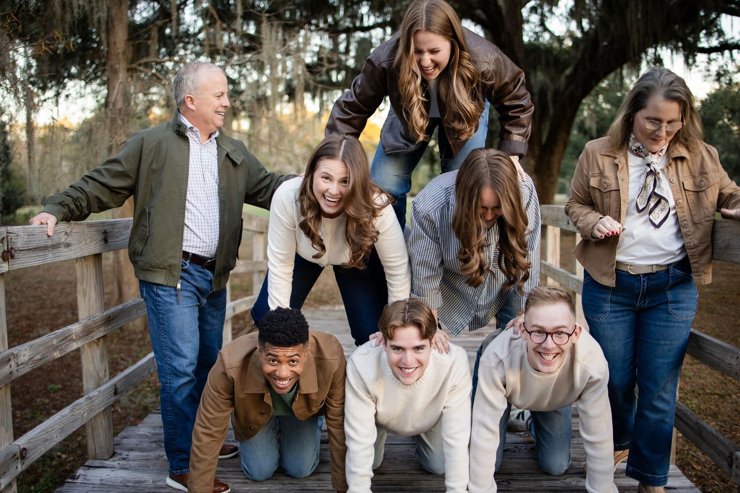 Group creates playful human pyramid formation on wooden bridge in park setting.