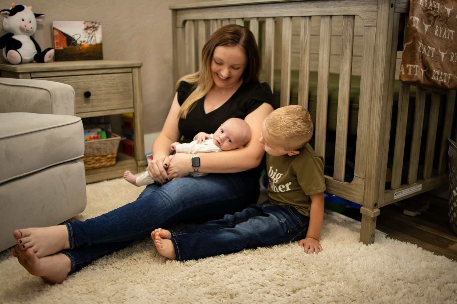 A nurturing moment captured in a cozy nursery as a parent in black shirt and jeans feeds a baby on a plush white carpet.
