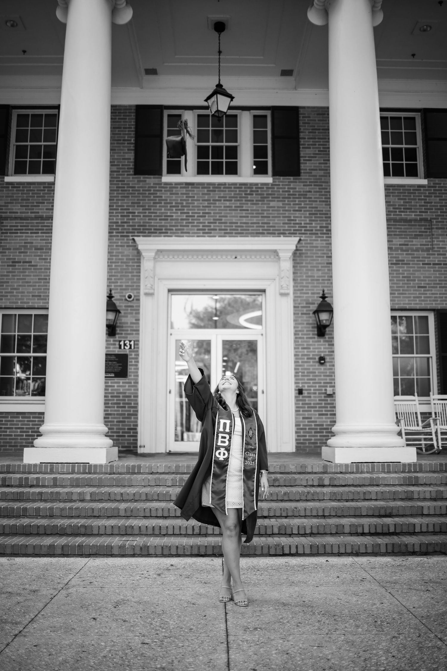 A graduate in academic regalia poses on the steps of a grand brick building with white columns in black and white.