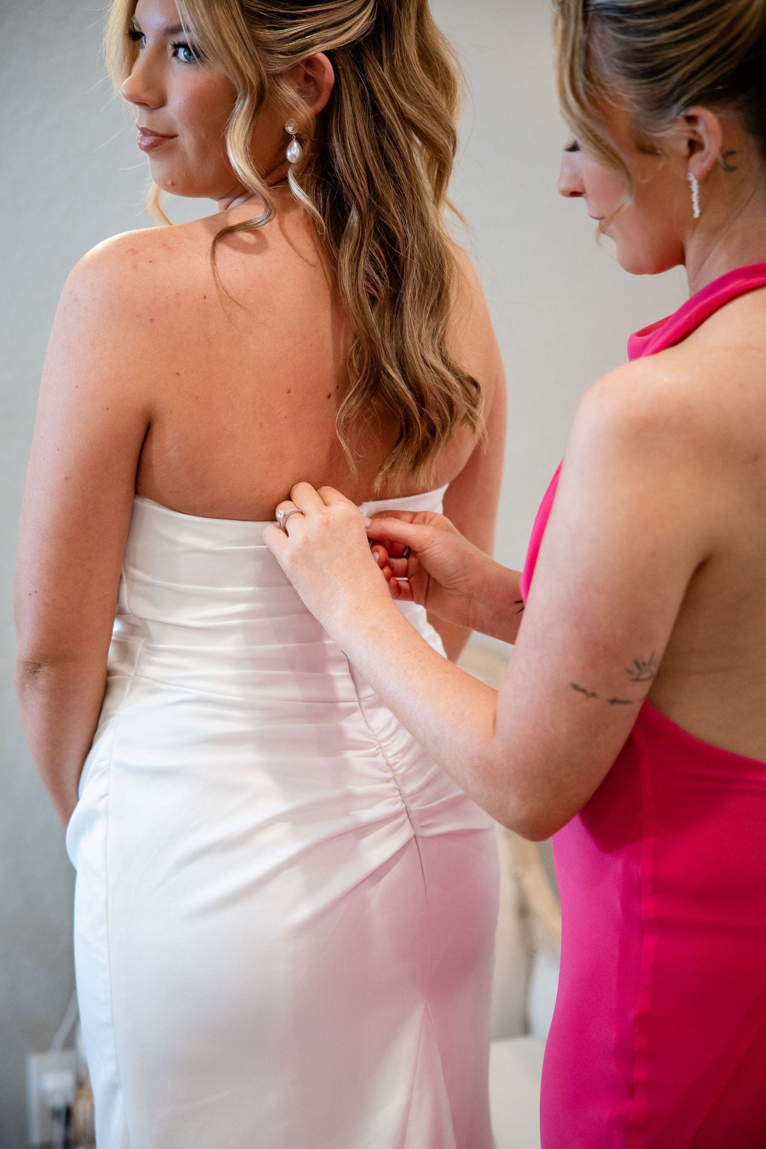 Bridesmaid in pink dress fastening buttons on back of bride's white strapless wedding gown.