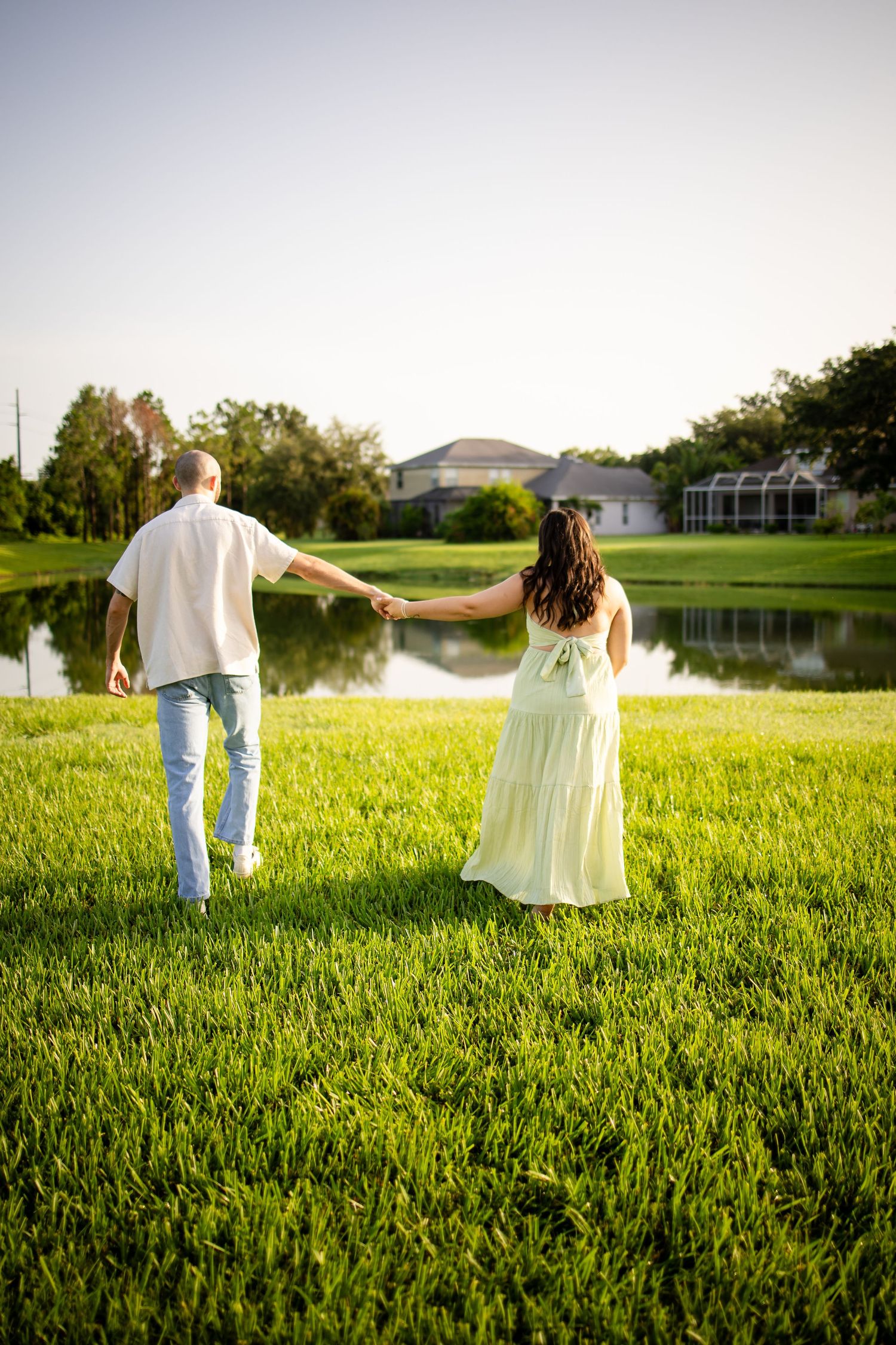A couple walks while holding hands in a lush green field near a lake during golden hour.