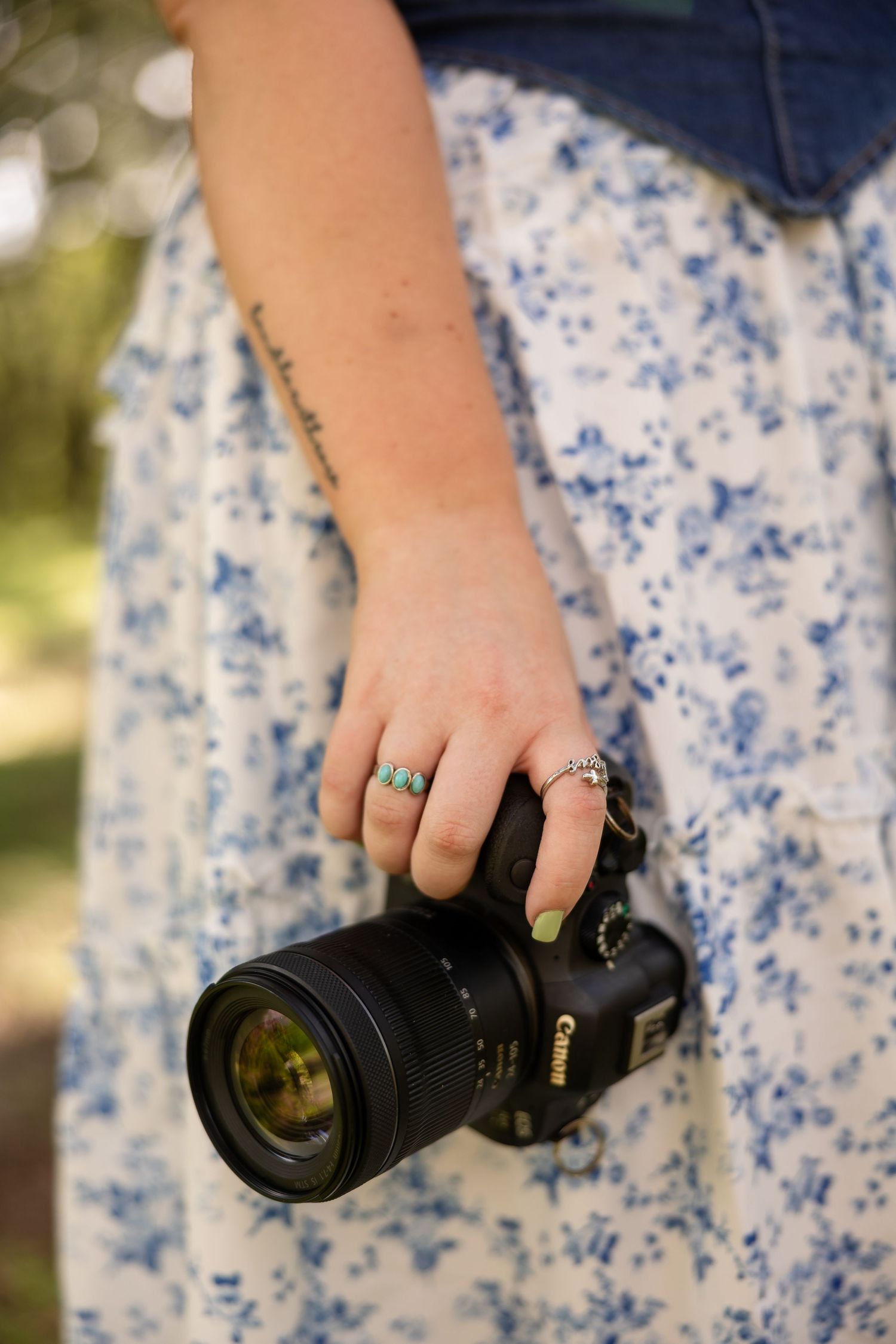 A hand wearing turquoise and silver rings holds a black DSLR camera against a blue and white floral dress.